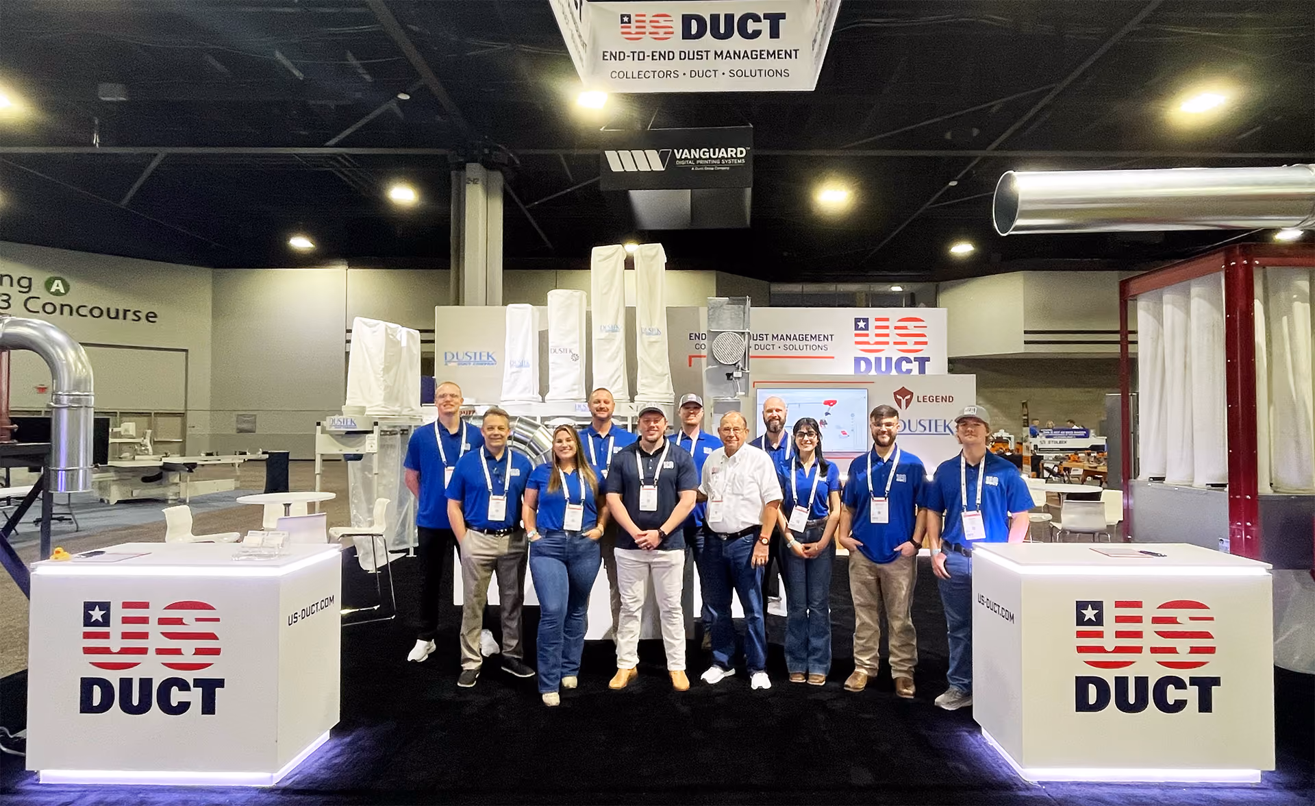 Group of eleven people in blue and white shirts posing at US Duct trade show booth with equipment.