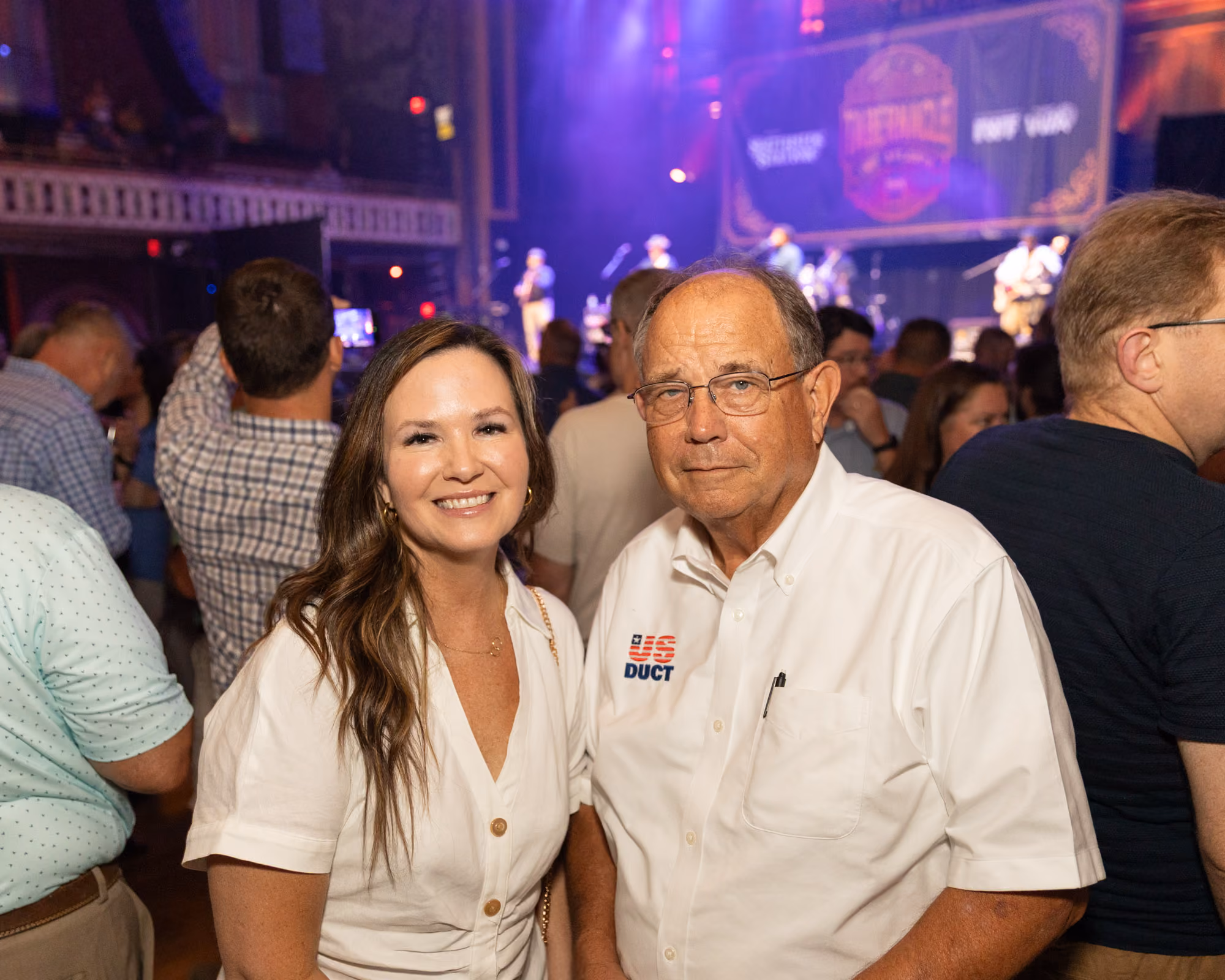 Smiling woman and man wearing glasses standing in a crowd at a live music event with a lit stage.