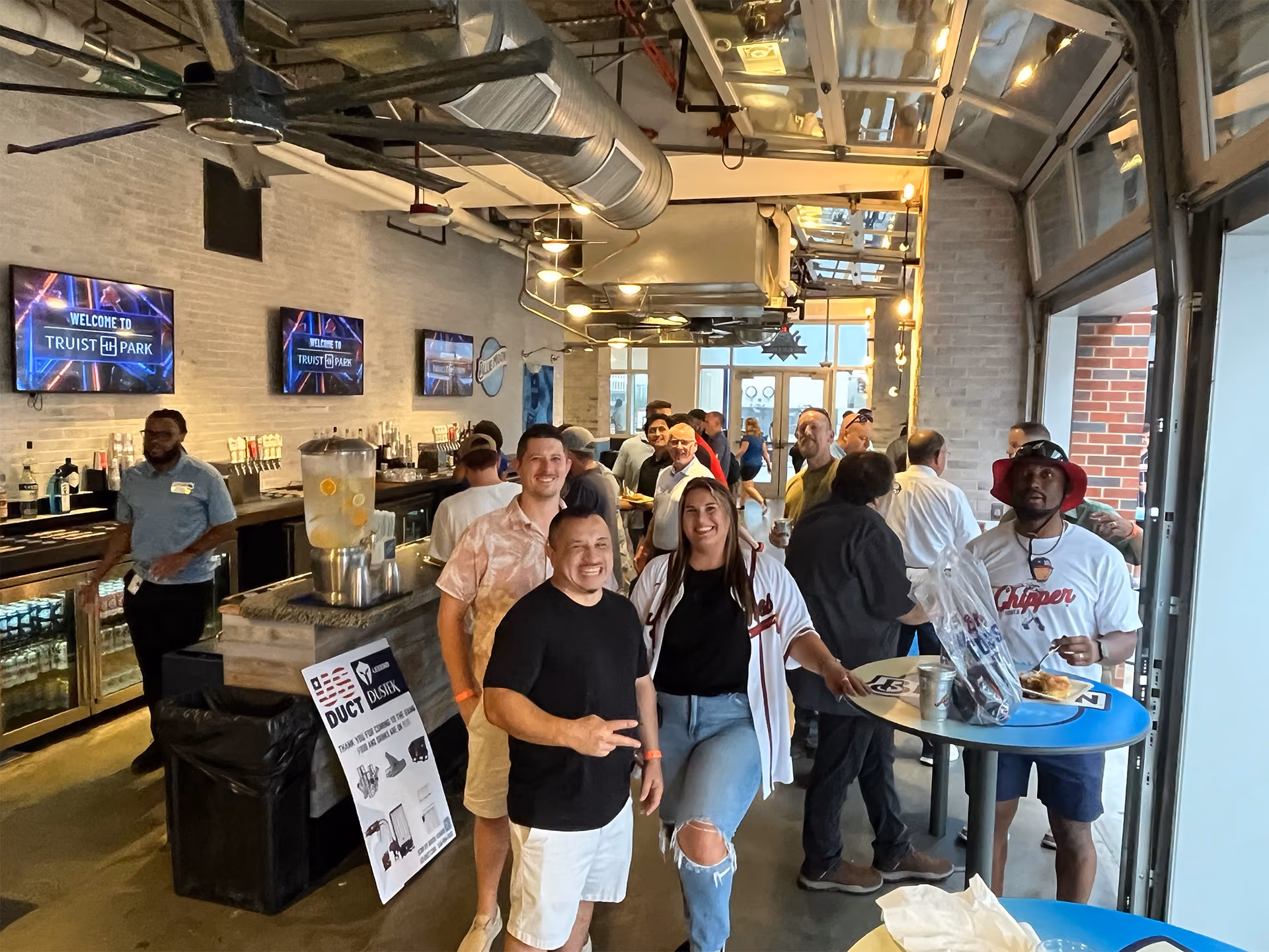 Group of people socializing and smiling at an indoor bar area with multiple TV screens.