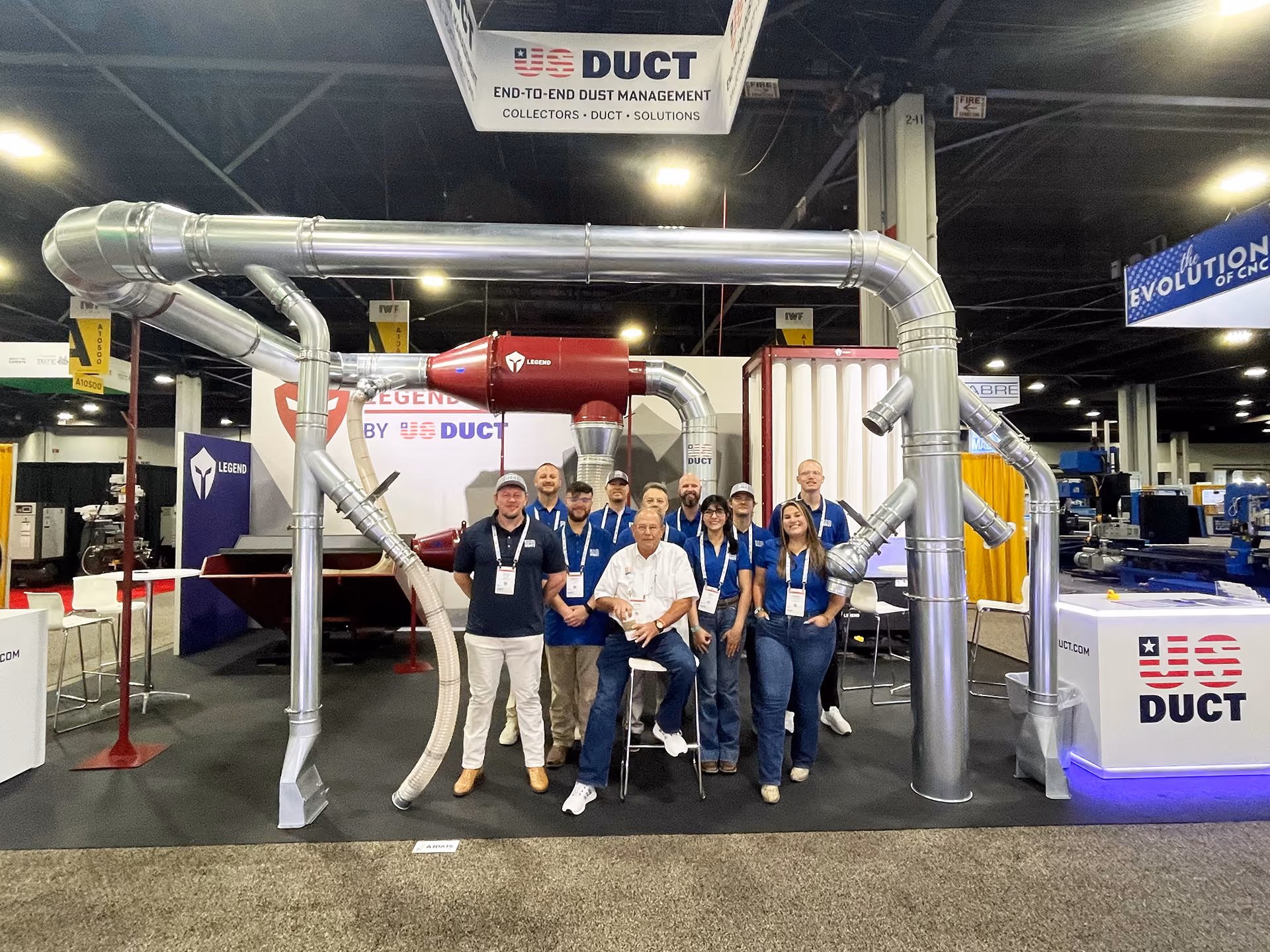 Group of nine people posing inside a trade show booth with large industrial ductwork and US DUCT branding.