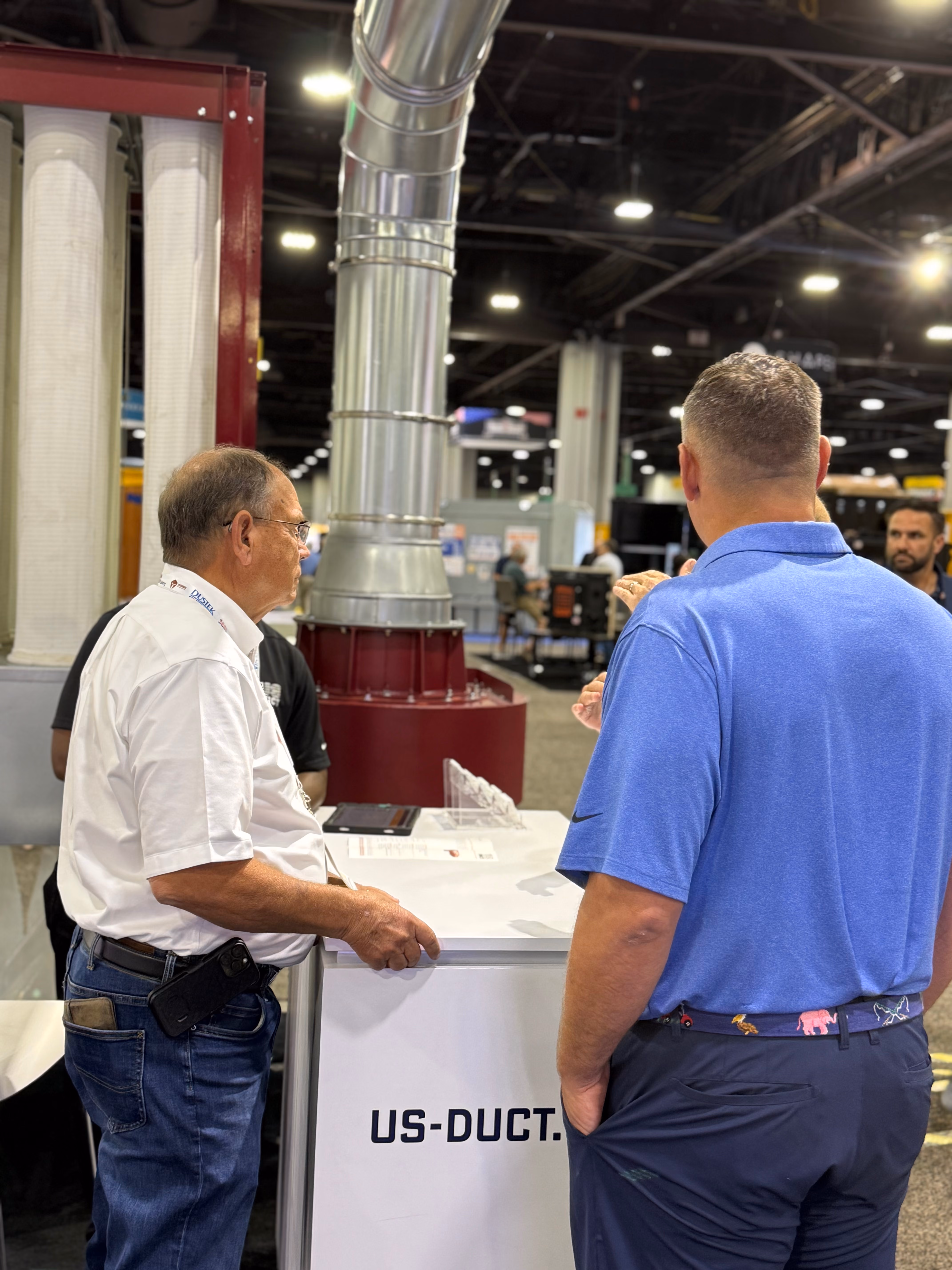 Two men talking at US-DUCT booth with ductwork display at an indoor trade show.