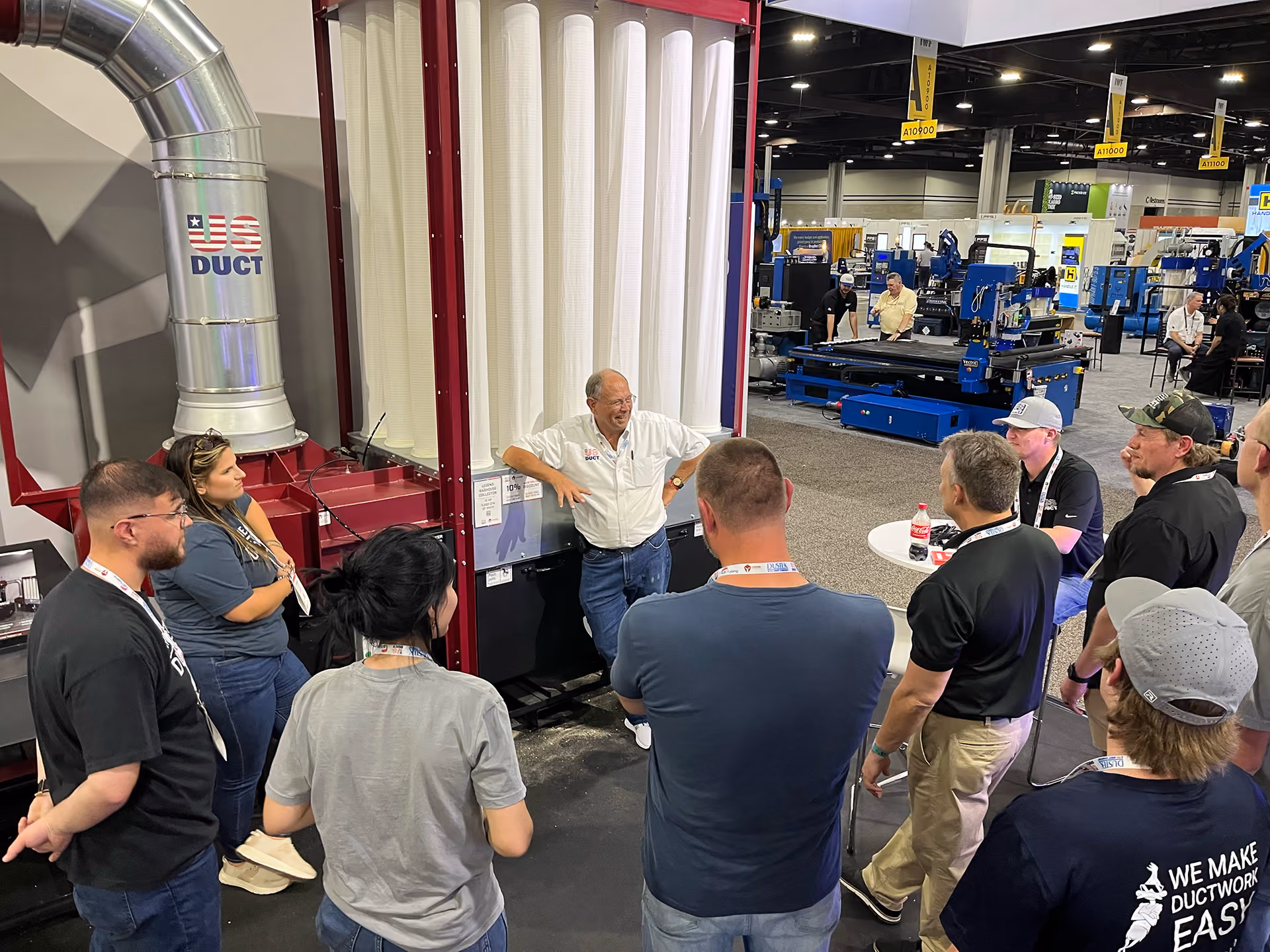 Man in white shirt talks to a group around industrial ductwork at a trade show booth.