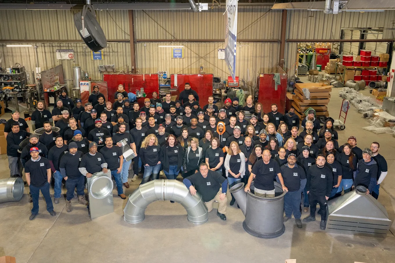 A large group of people, all wearing matching black long-sleeve shirts with a white logo, pose inside a spacious industrial warehouse with metal ductwork pieces arranged in front of them on the floor.