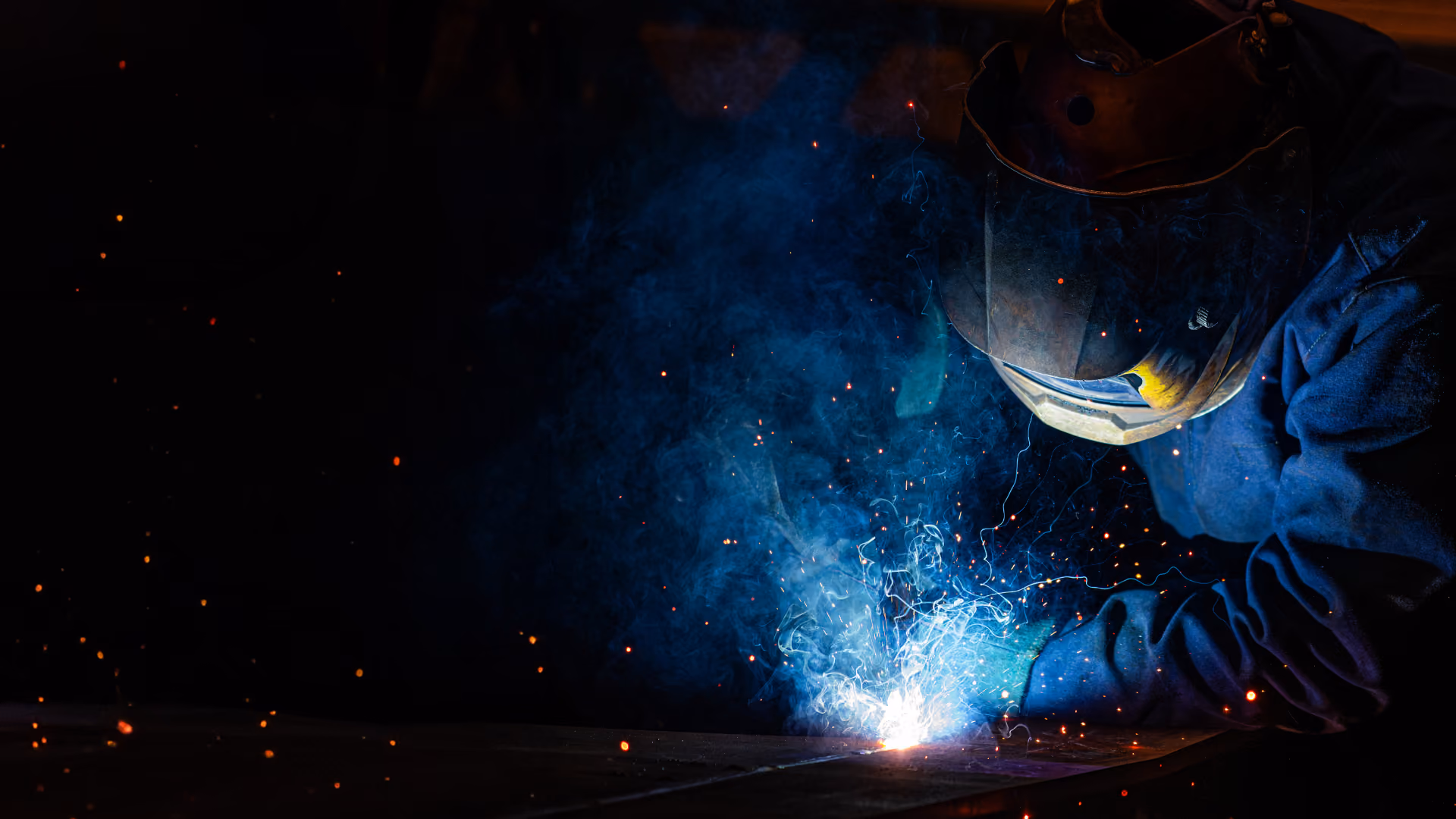 Person wearing protective welding helmet and gloves welding metal with bright blue sparks and smoke in a dark environment.