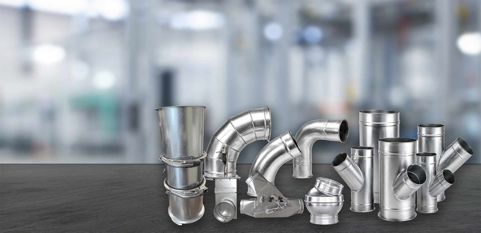 A collection of various shiny metal HVAC duct components including straight pipes, elbows, tees, and connectors arranged on a dark surface with a blurred industrial background.