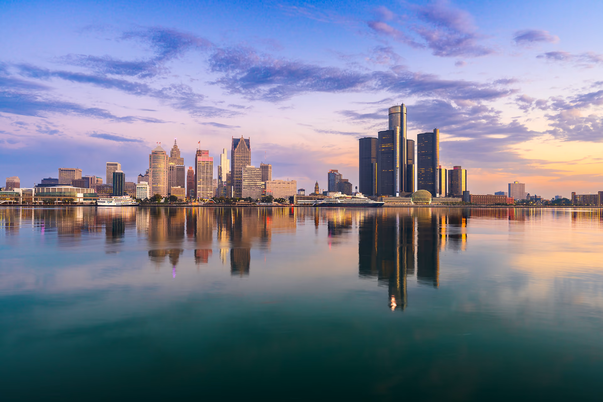 Sunset view of Detroit skyline with city buildings and water reflecting the sky and lights.