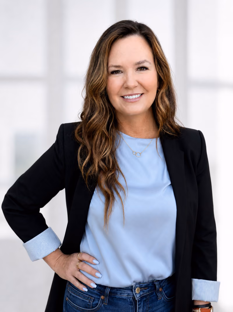 Smiling woman with long wavy brown hair wearing a light blue blouse, dark blazer with rolled cuffs, and jeans, standing with one hand on her hip against a light blurred background.