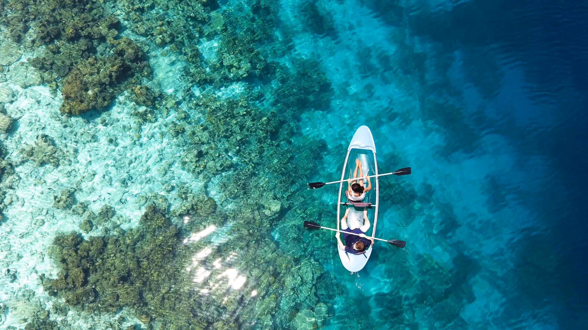 Aerial view of two people paddling a clear, transparent kayak over vibrant turquoise water with visible coral reefs beneath.