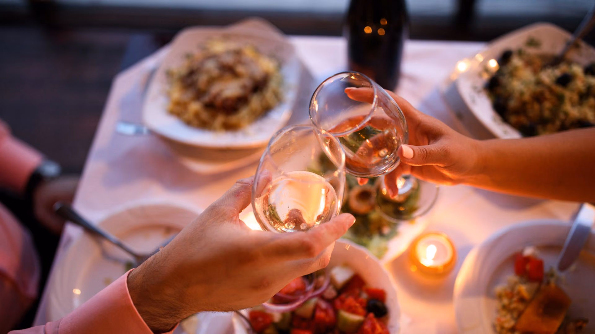 Two people clinking glasses of white wine over a table set with plates of food including salad, pasta, and a lit candle.