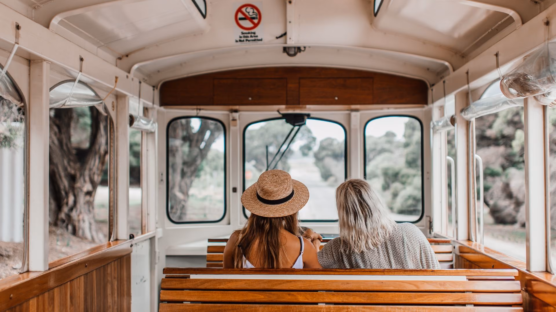 Two women sitting on wooden bench seats inside a vehicle facing front windows with trees visible outside, one wearing a straw hat and the other with blonde hair.