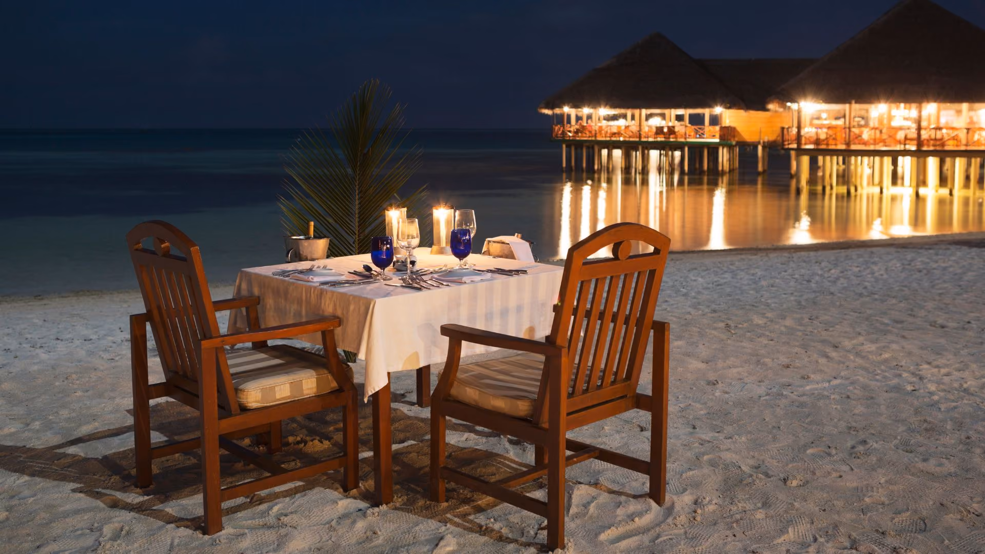 Two wooden chairs with cushions at a table set for dinner on a sandy beach at night, with lit candles, blue glasses, and a bottle of wine in an ice bucket, and illuminated overwater bungalows reflected in the calm water in the background.