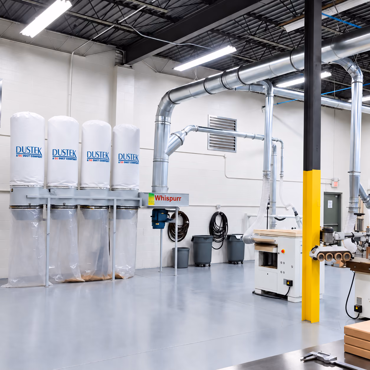 Interior of a clean, well-lit industrial workshop with dust collection equipment labeled DUSTEK and Whispurr, connected by large metal ducts, along with wood processing machines and organized storage bins against white walls and a gray floor.