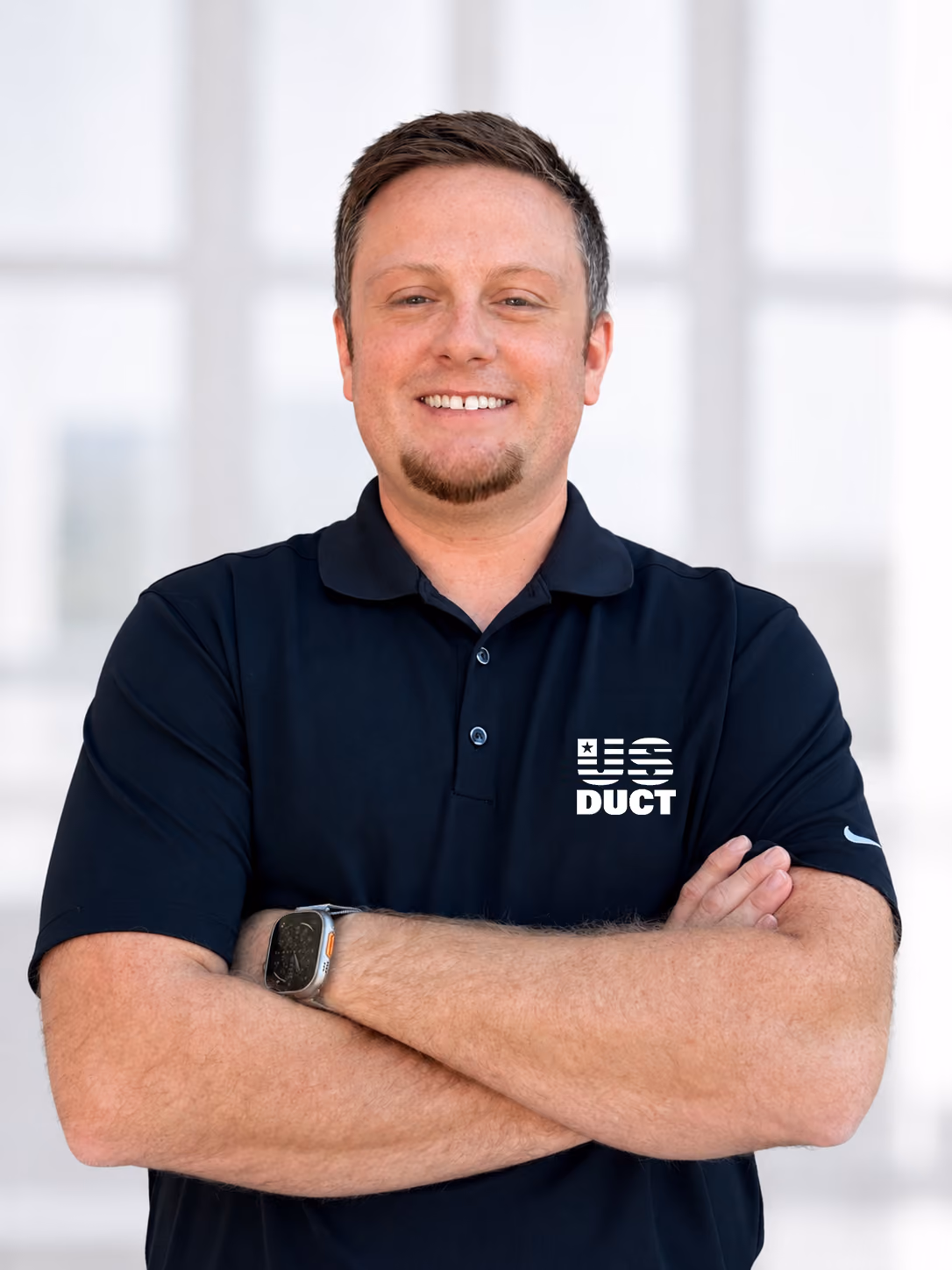 Man with short brown hair and a goatee smiling with arms crossed, wearing a black polo shirt with a white US DUCT logo on the chest and a smartwatch on his left wrist.