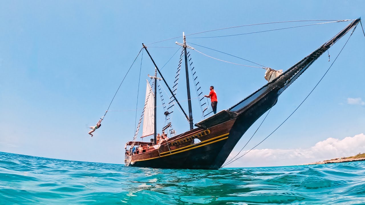 A wooden sailing ship named Jolly Pirates on bright blue water under clear sky with a person swinging on a rope from the ship and another standing on the ship's bow.