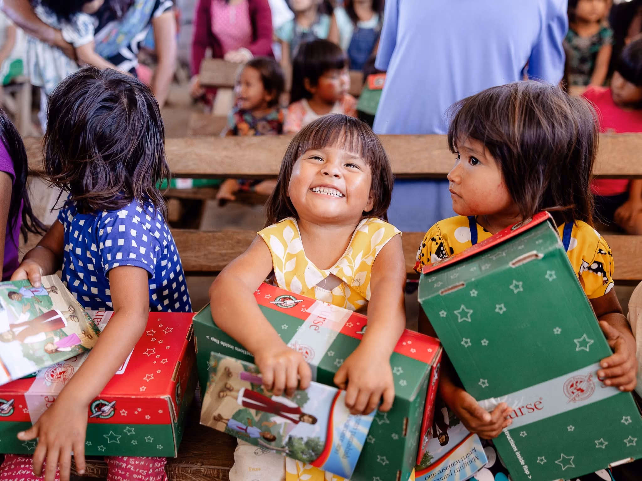 Three young children sitting on a wooden bench, each holding a green and red shoebox and a booklet, with one child in the center smiling brightly.