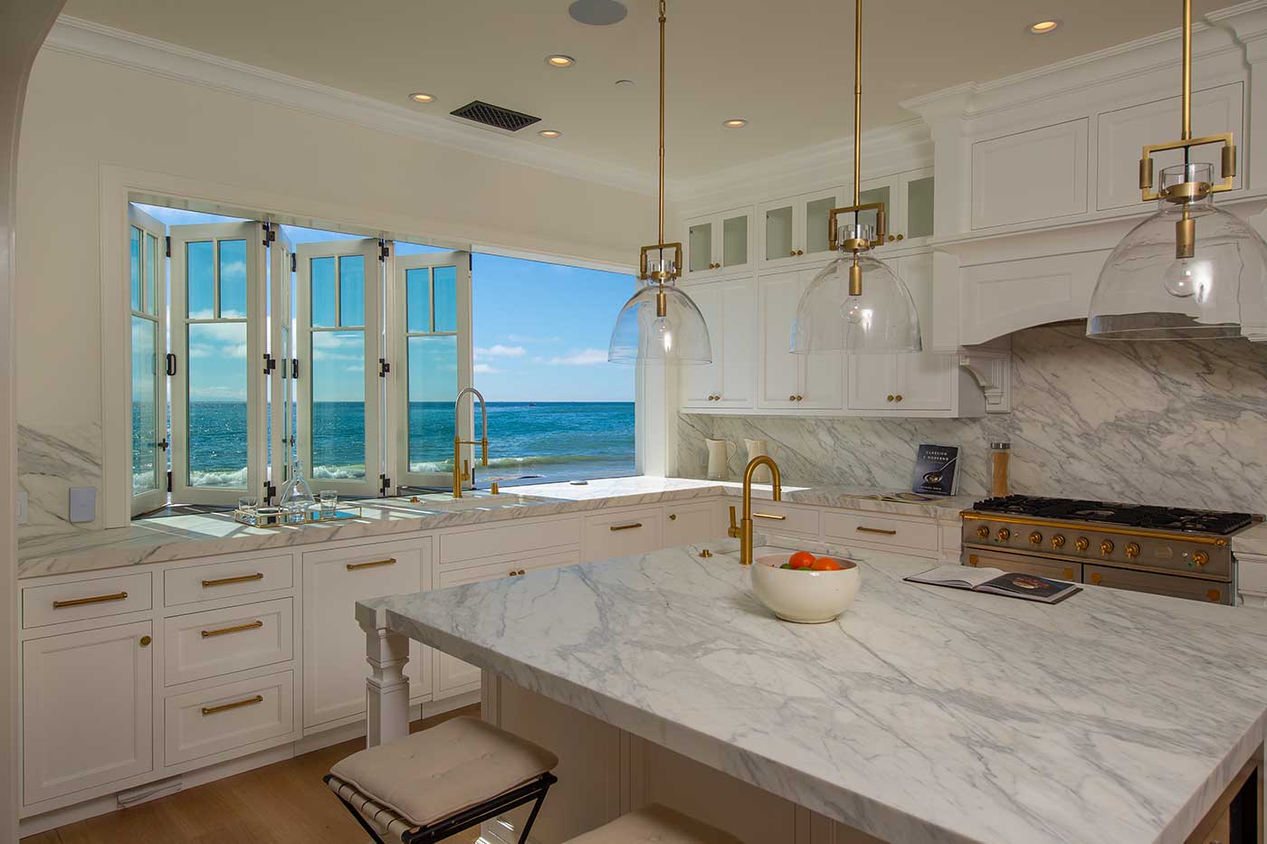 The 6-panel white bifold passthrough window with cottage-style divided lites, ocean views visible through the opening. Kitchen interior is white farmhouse style.