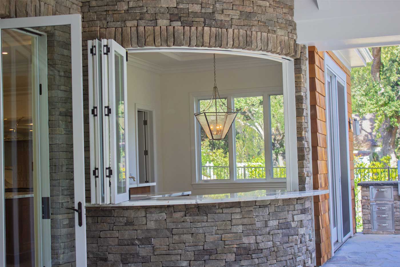 Tall, narrow radius-top bifold passthrough window framed by stonework. Interior wet bar visible, outdoor covered patio on the other side.