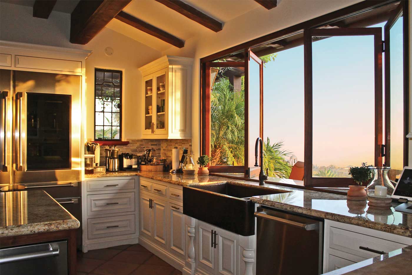 Dark-stained wood bifold passthrough window in a Spanish farmhouse kitchen. Warm interior lighting, Santa Barbara landscape visible through the opening.