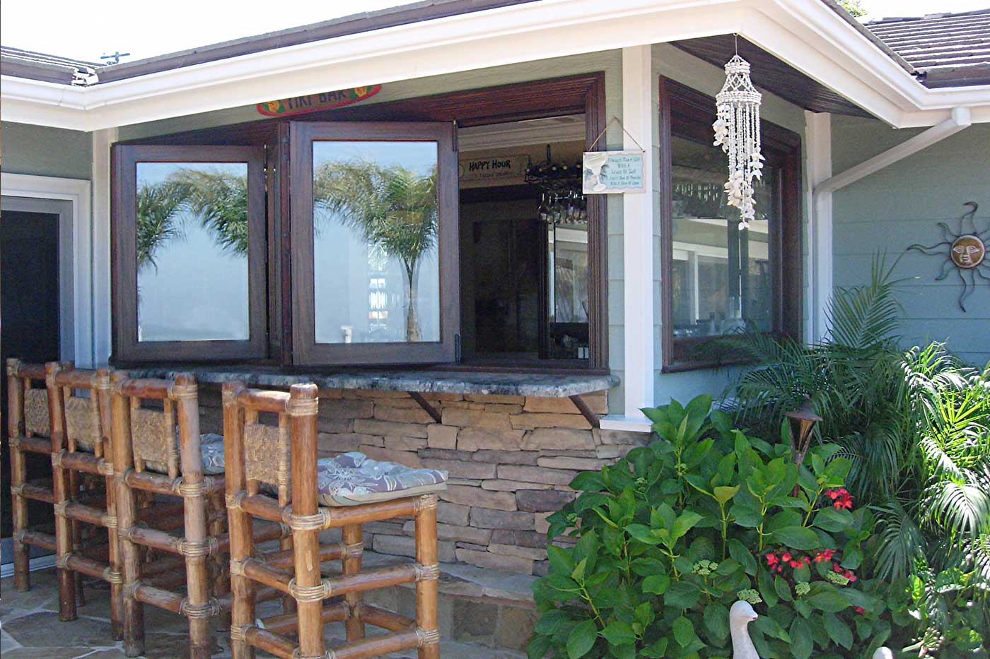 Tropical backyard tiki bar with 4-panel all-wood bifold passthrough window. Fully stocked bar inside, pool and tropical landscaping visible outside. Stonework base.