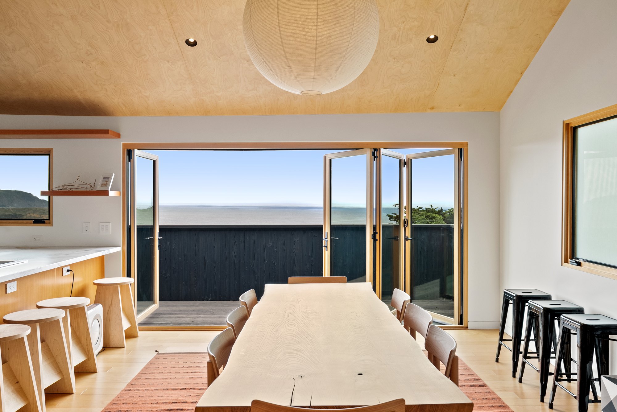 Wood bi-fold doors folded open at both sides of a kitchen and dining room, with a long wood table leading out to an ocean-view deck.