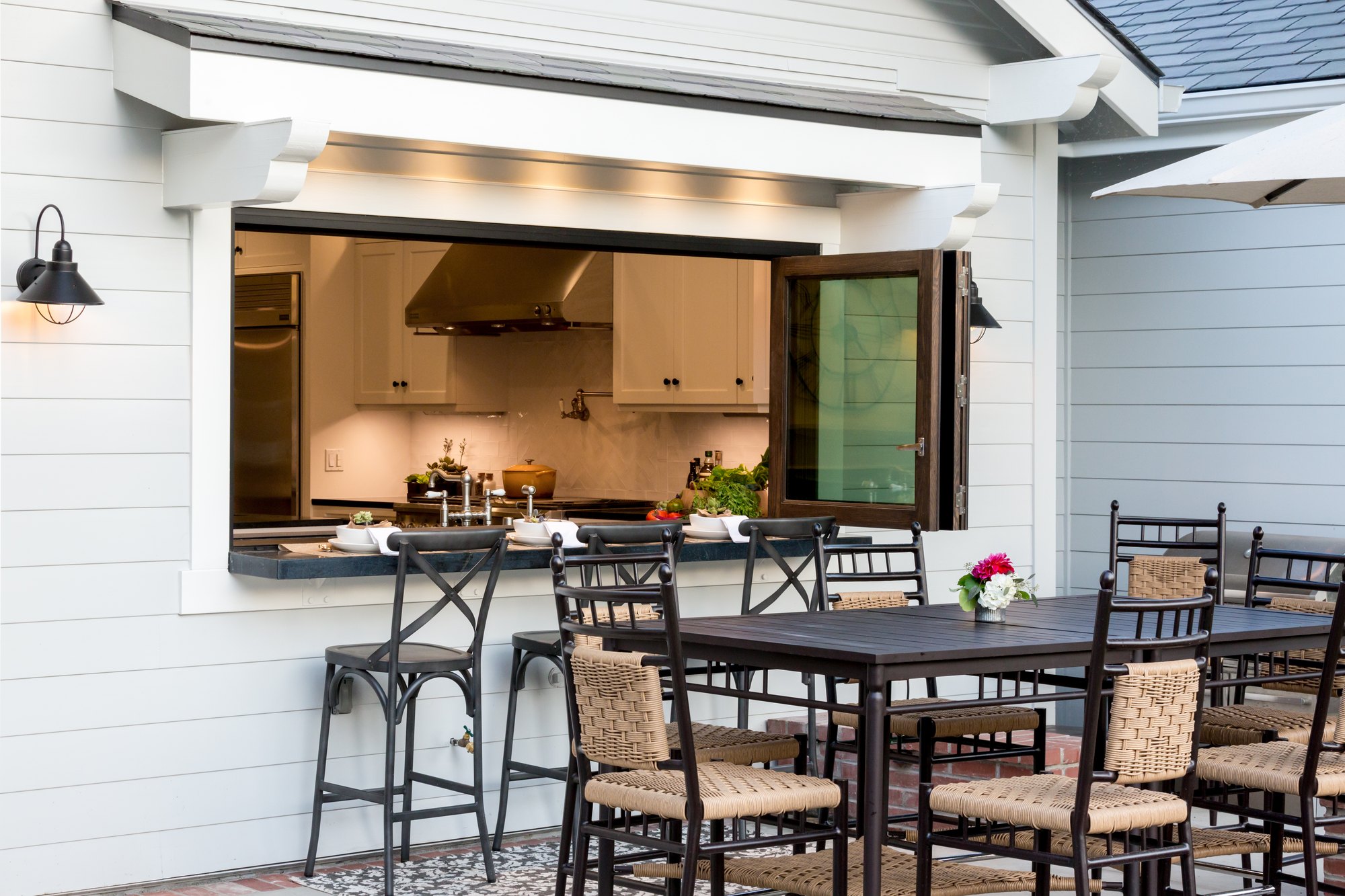 Bi-fold kitchen pass-through window folded open above an exterior bar counter, with bar stools and a patio dining table outside. 