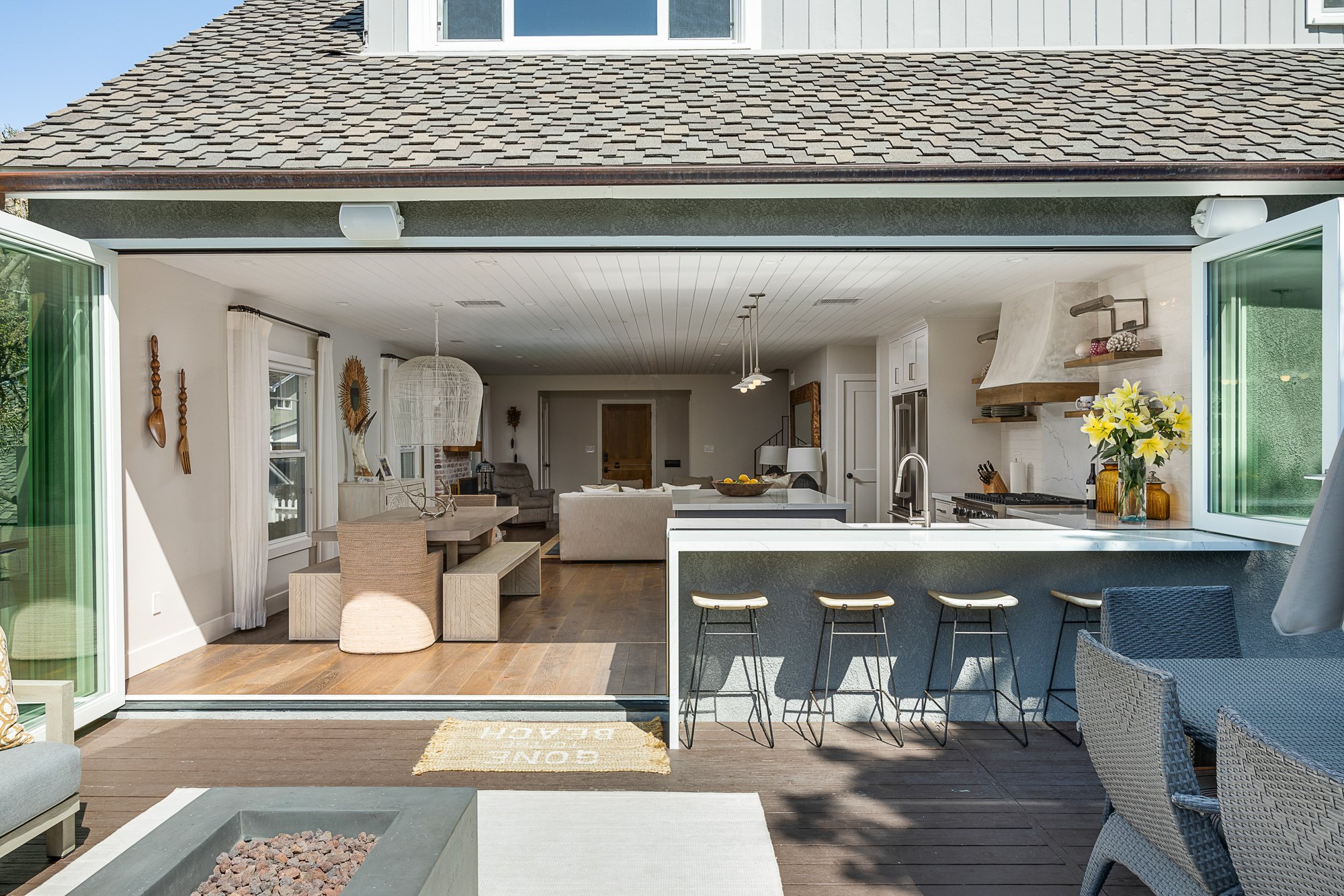 Bi-fold window and bi-fold door folded open together, removing the kitchen's back wall and turning the counter into an outdoor bar with stools on the deck.