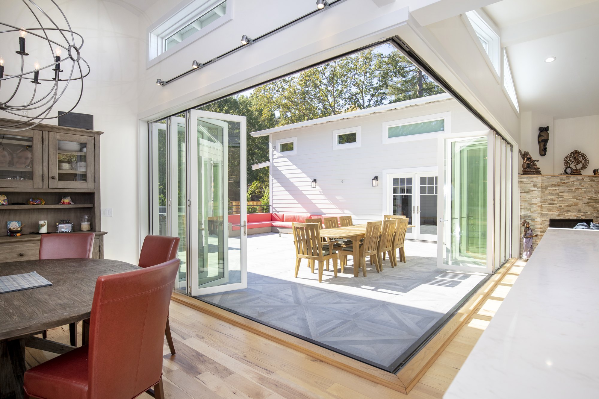 Zero-corner bi-fold doors folded open at a 90-degree corner with no post, connecting a dining table to a courtyard with outdoor dining. 