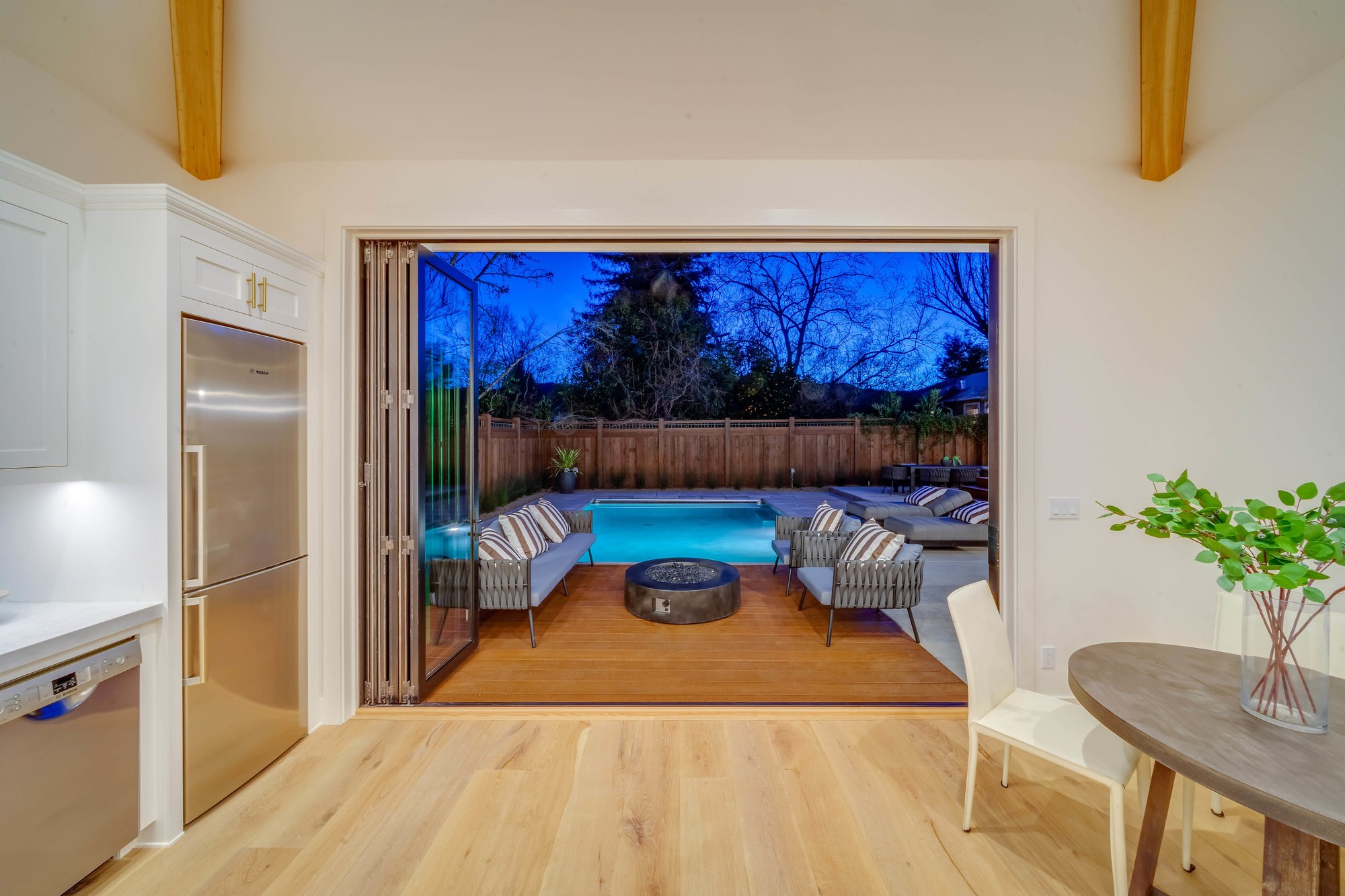 Kitchen at dusk with bi-fold doors folded open to a lit pool deck with lounge seating and a fire bowl.