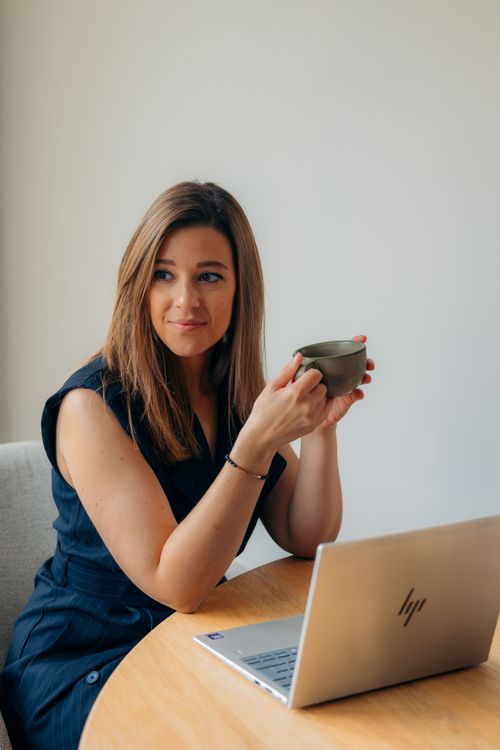 Laura Gielis zit in een zakelijke setting ontspannen met een kop koffie in de hand – uitnodiging tot persoonlijk contact en begeleiding op maat voor bedrijven.