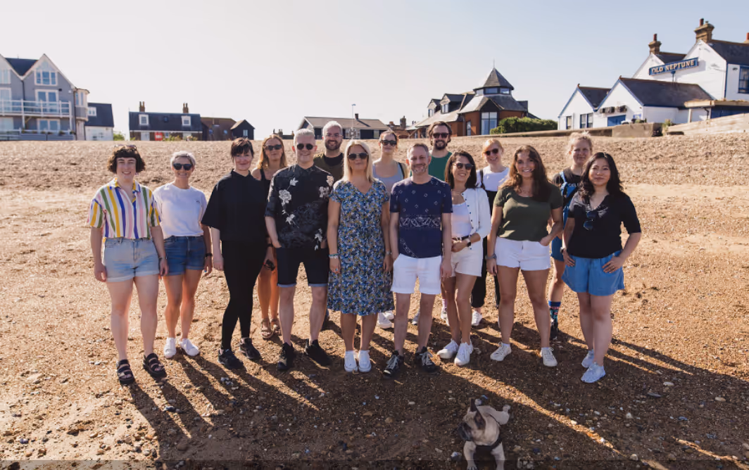Group photo of team members standing on the beach in Whitstable, England, with The Old Neptune pub and seaside houses in the background, under clear blue sky with sunshine. French bulldog dog in foreground.