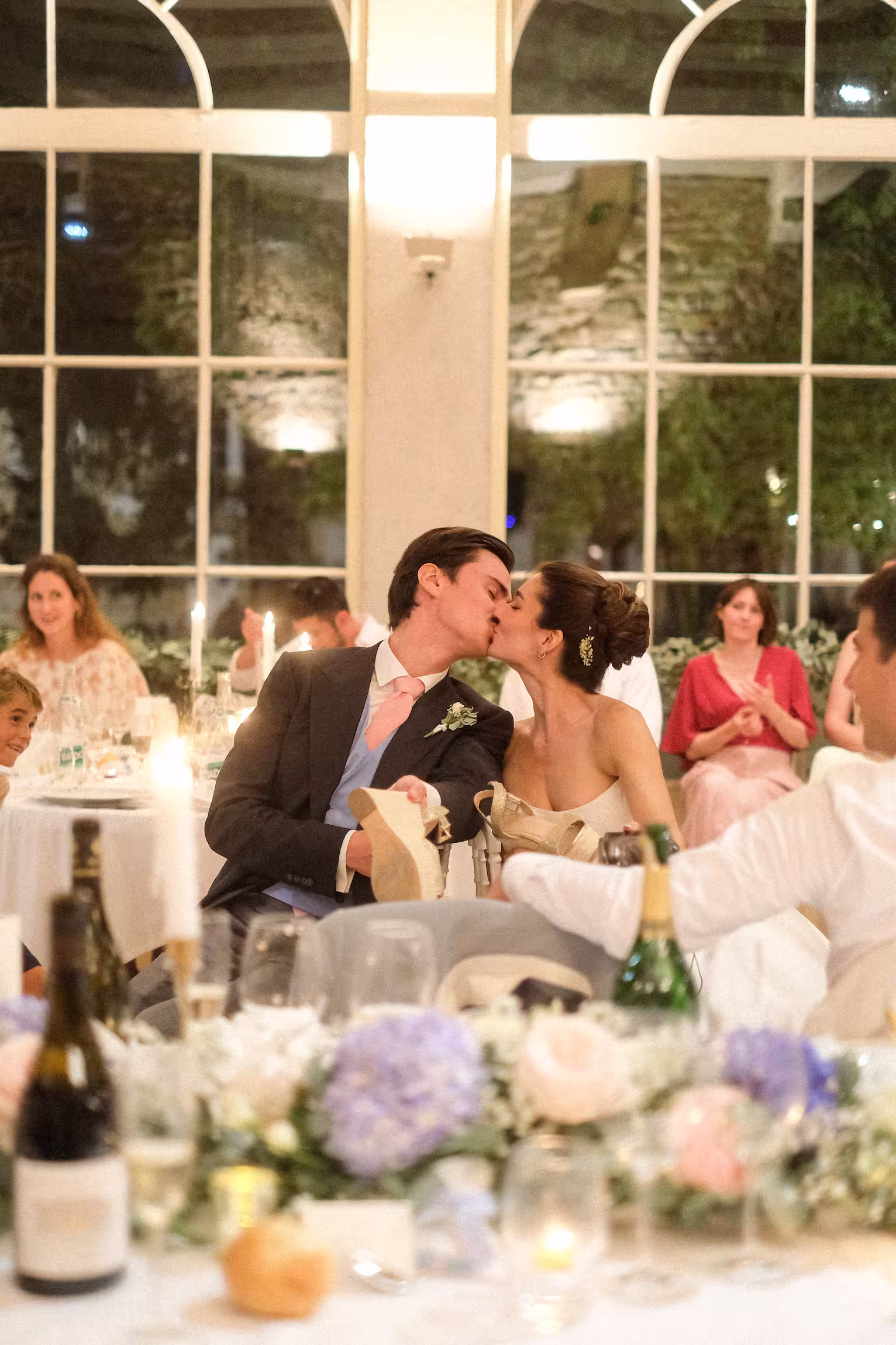 Les mariés s’embrassent à table pendant le dîner, entourés de fleurs et de bougies.