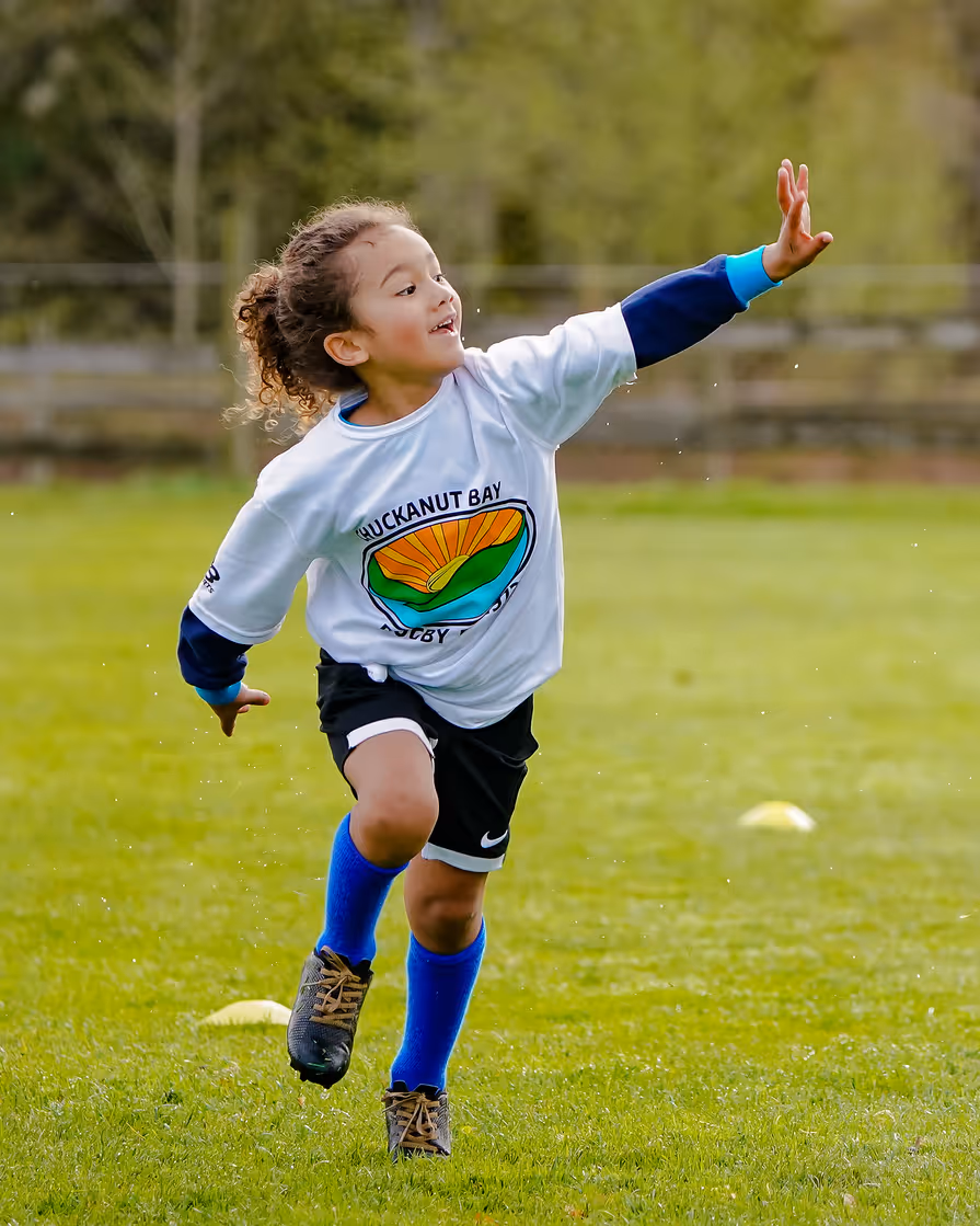 Young child playing rugby on grass field wearing Chuckanut Bay Rugby Club shirt with arm extended forward.