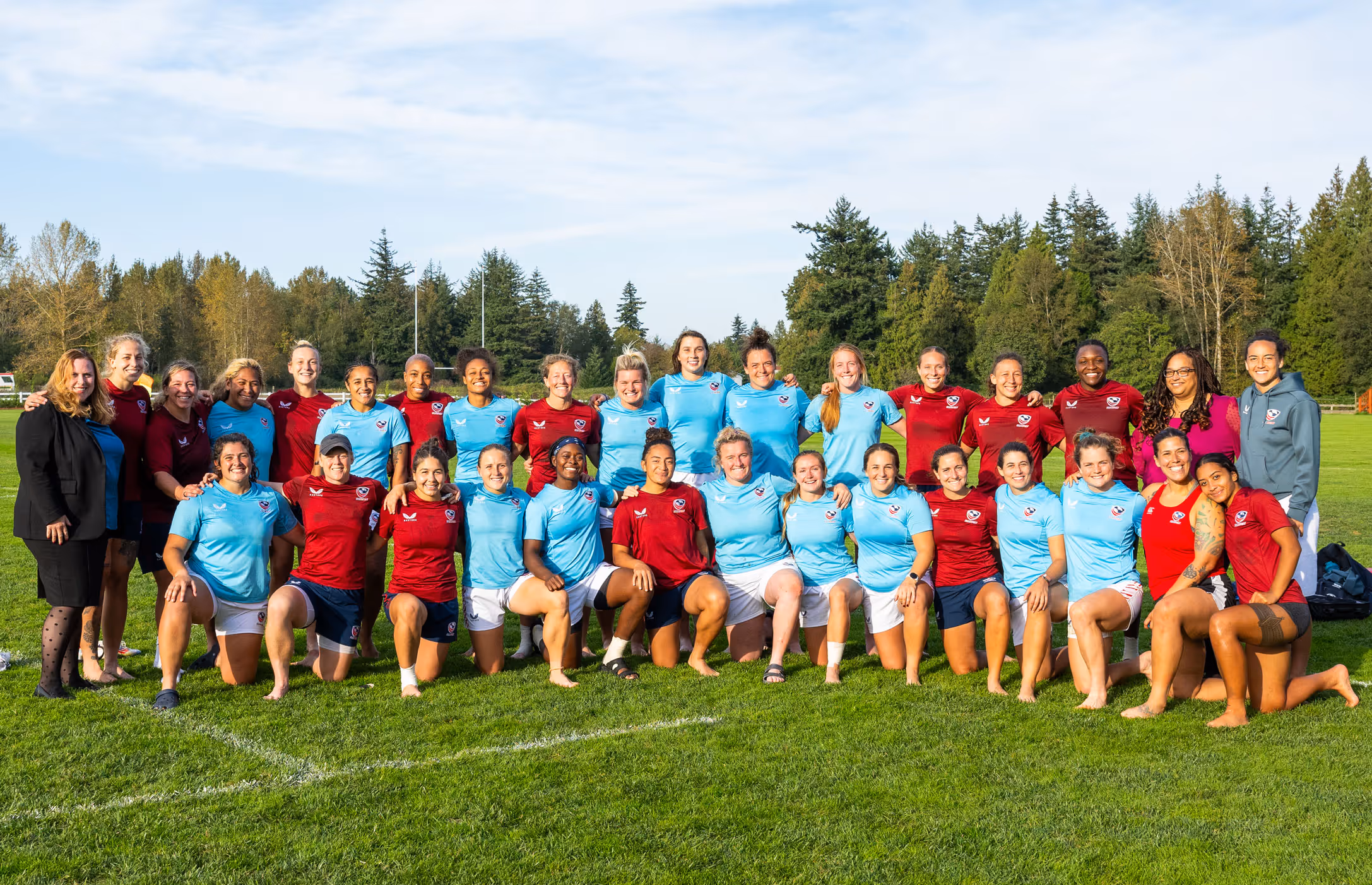 Large women's rugby team posing together on green field, some wearing red jerseys and others in light blue, with trees and blue sky in the background.