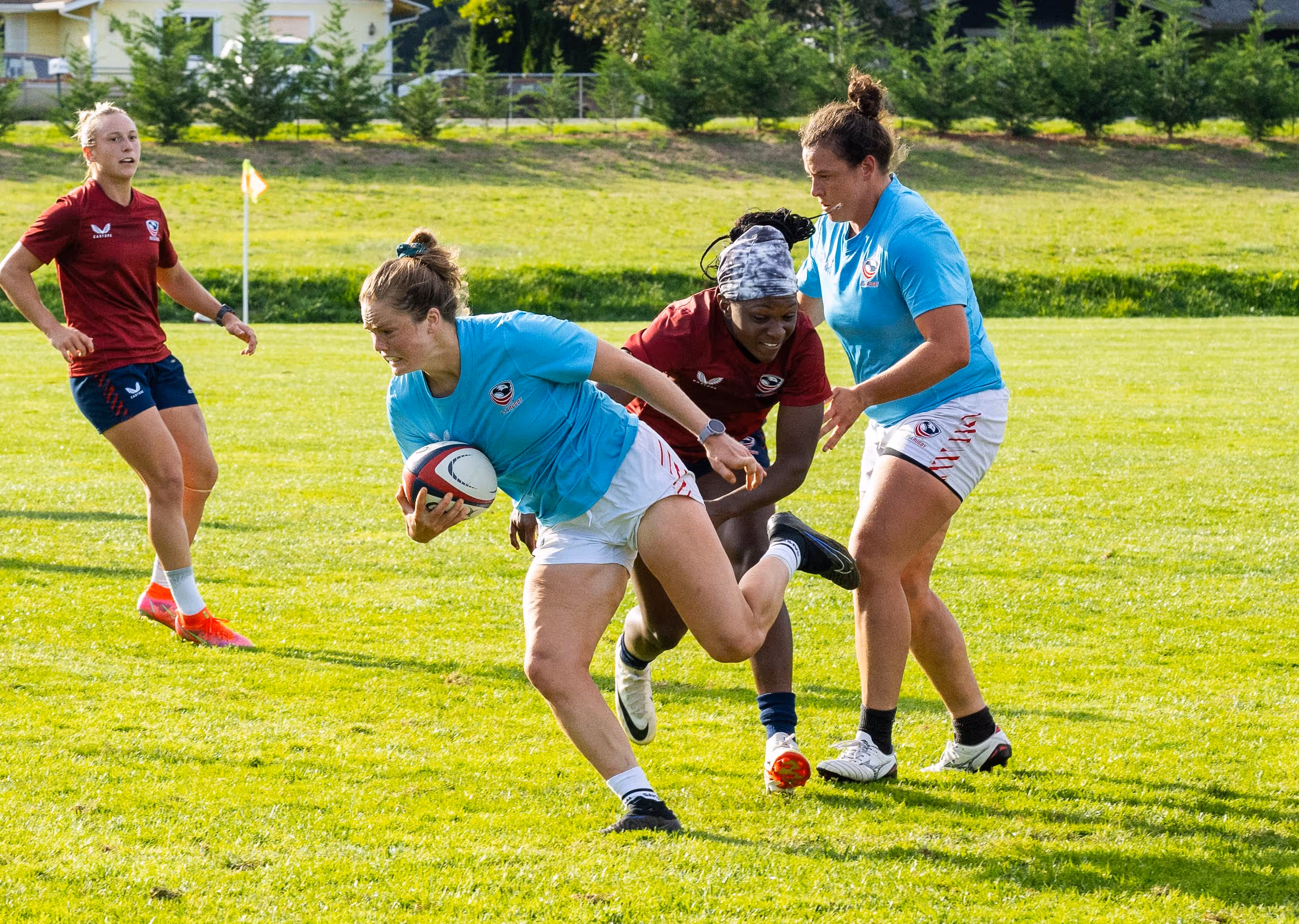 Female rugby player in light blue jersey running with ball while being tackled by opponents in red jerseys on grassy field.