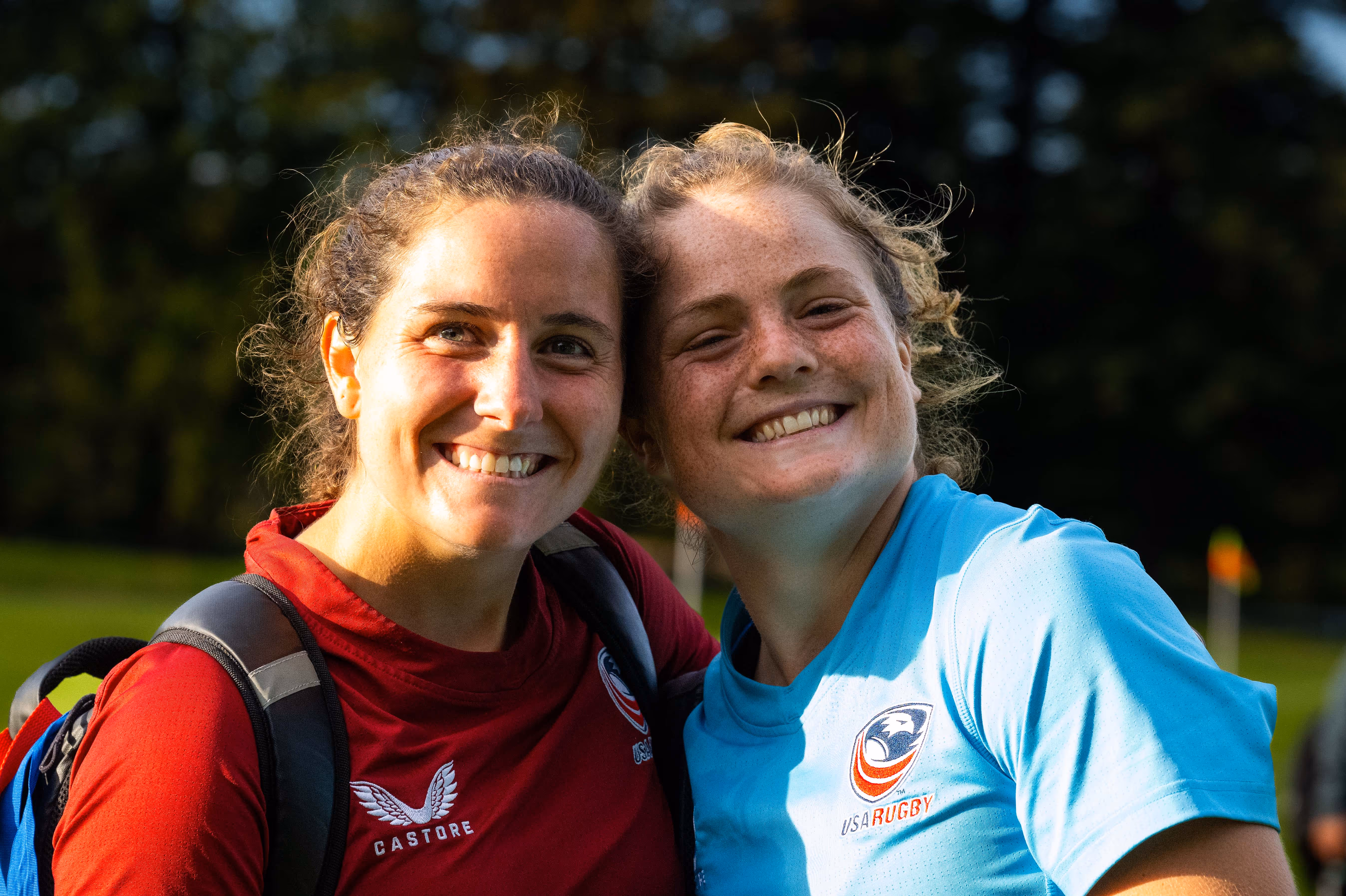Two smiling female rugby players wearing USA Rugby jerseys, one in red and one in light blue, posing outdoors on a sunny day.