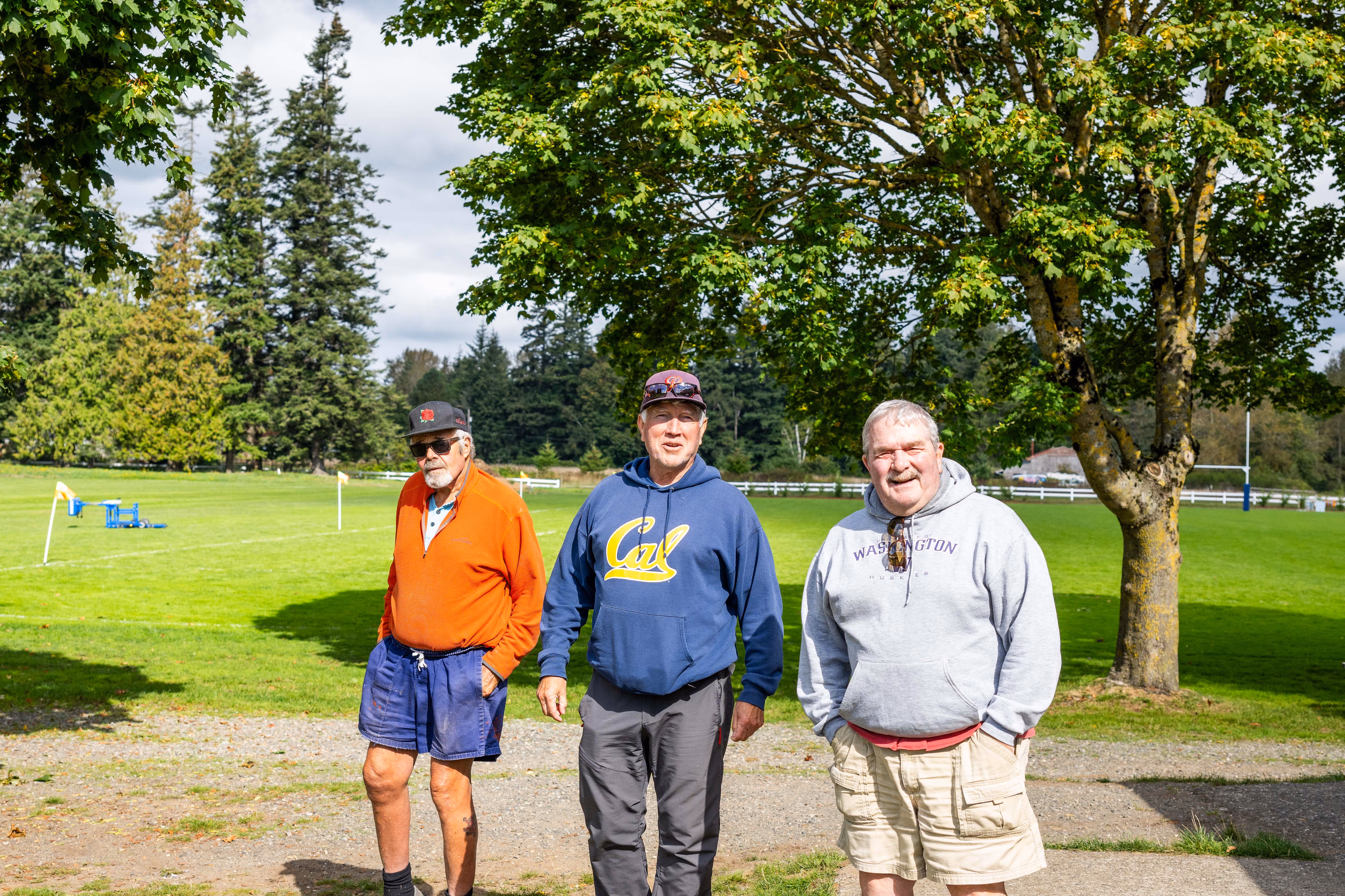 Three older men standing outdoors on a sunny day with trees and a grassy field behind them.