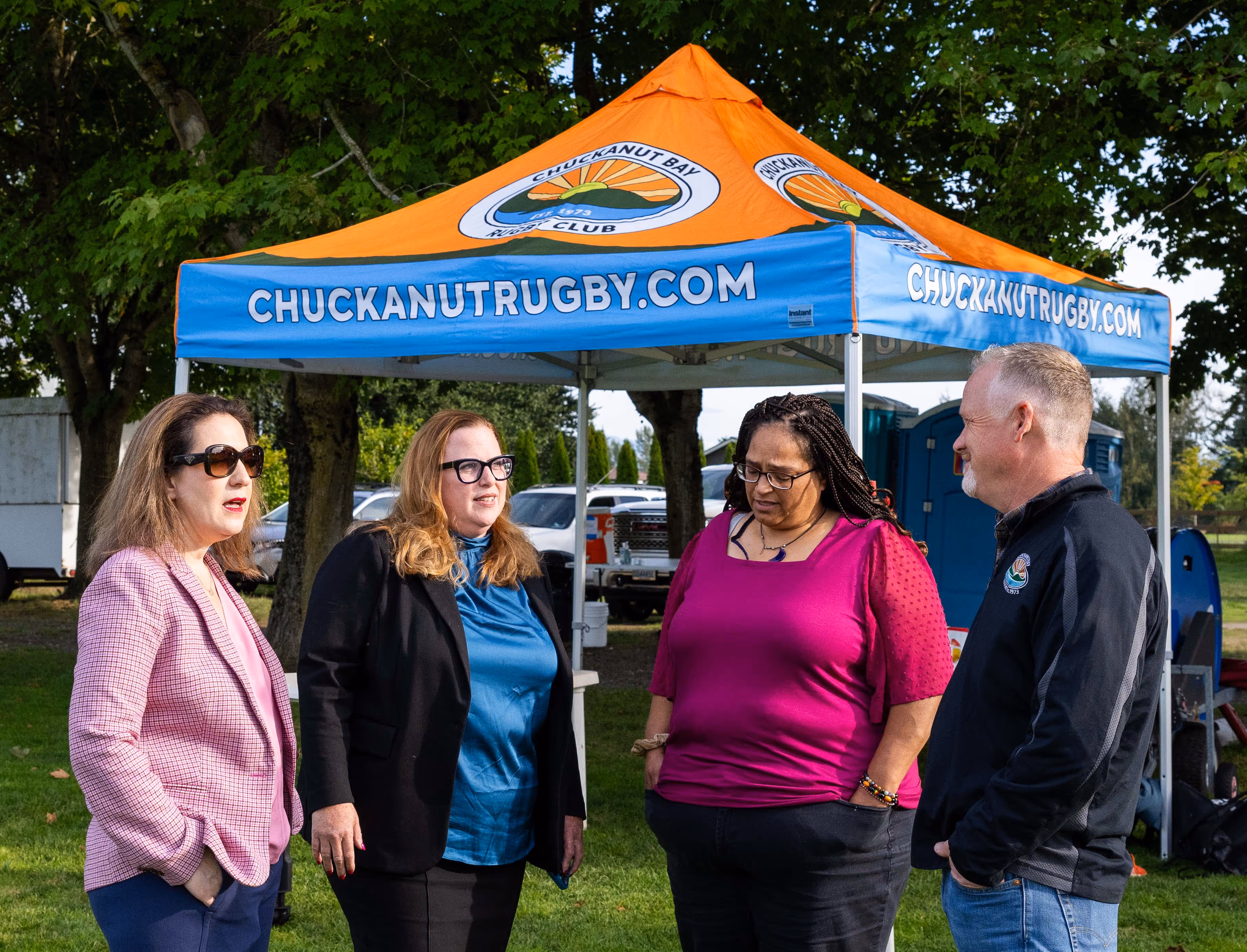 Four adults conversing outdoors in front of an orange and blue tent with 'CHUCKANUTRUGBY.COM' on its sides.