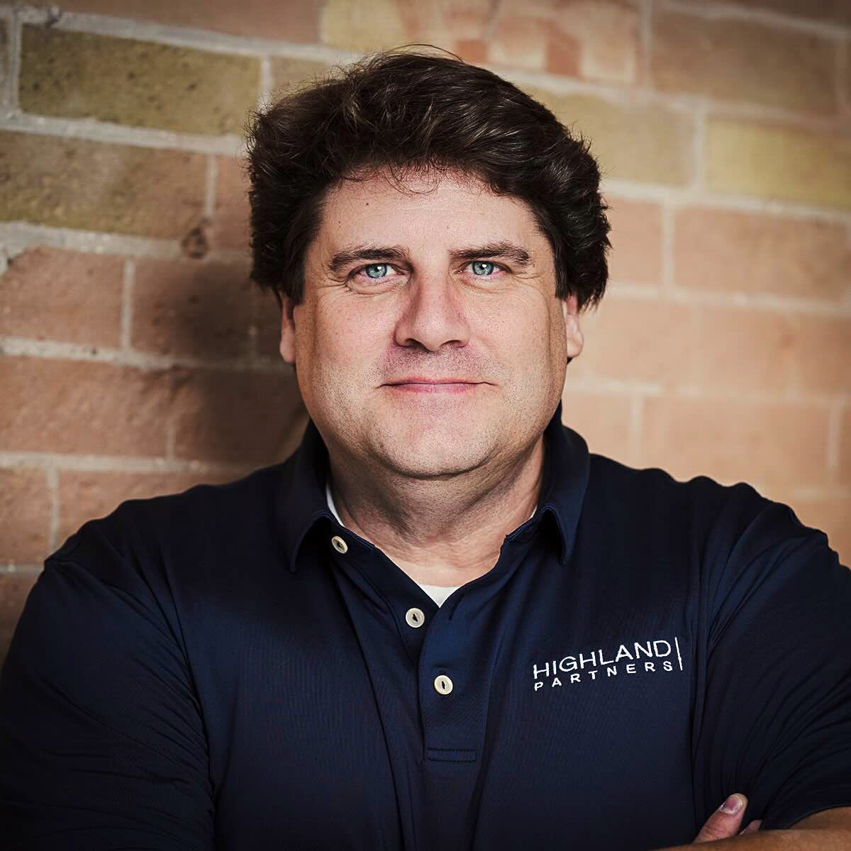 Man with dark hair and blue eyes wearing a navy polo shirt with Highland Partners logo, standing against a brick wall.