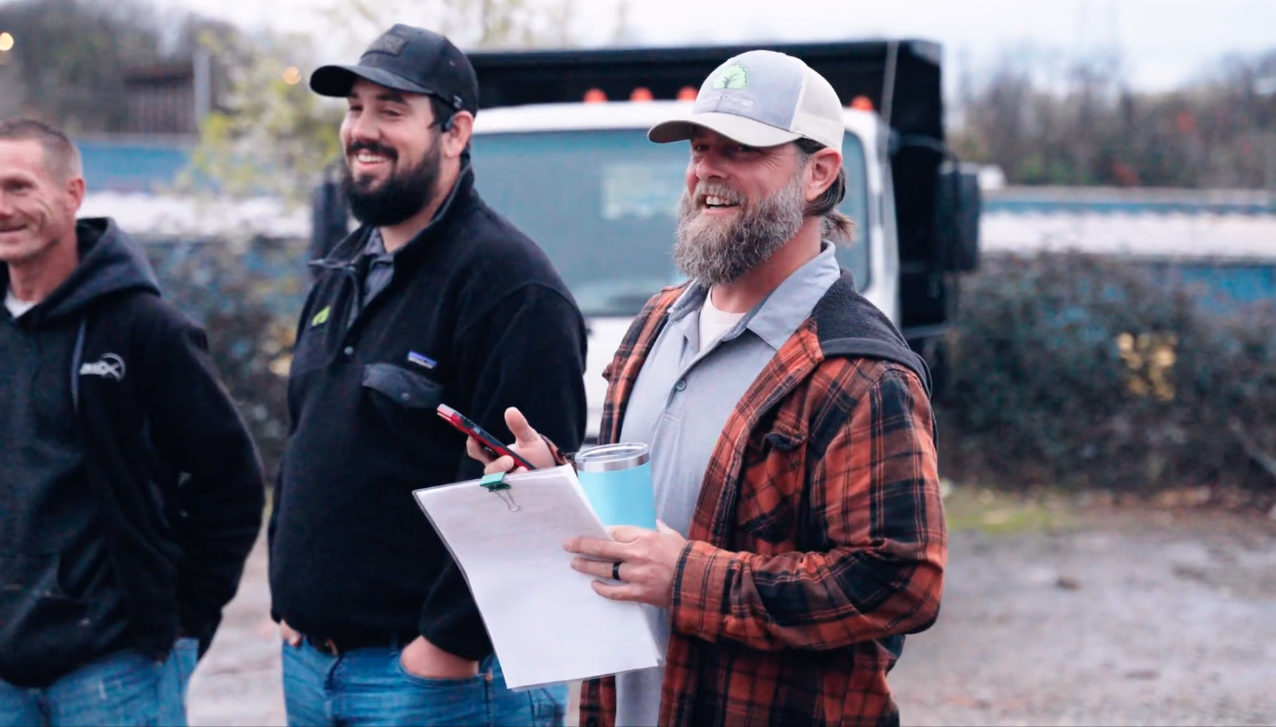 Three men in casual outdoor attire stand near a dump truck, one holding a clipboard and a blue tumbler.