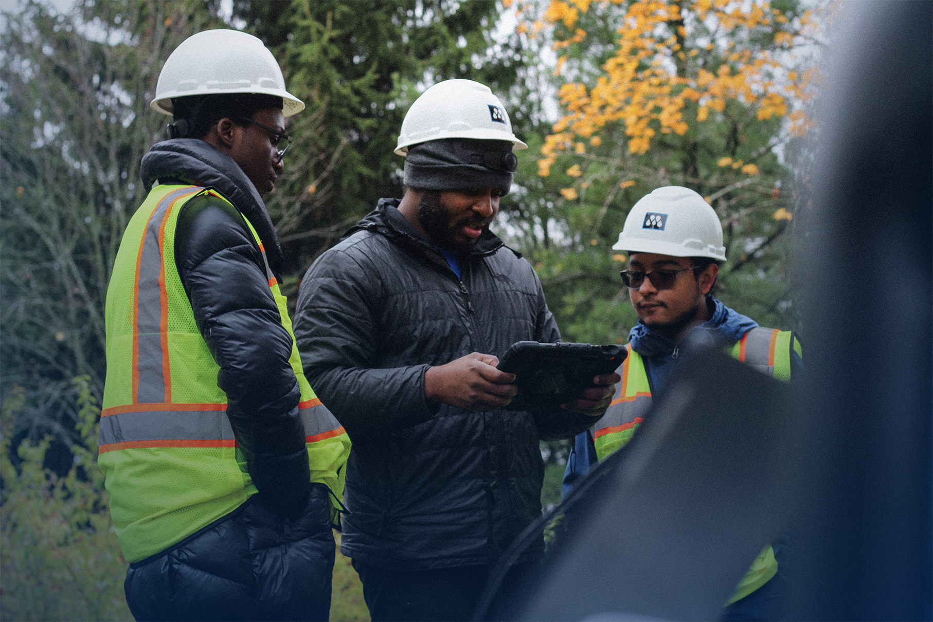 Three construction workers in safety vests and white helmets looking at a tablet outdoors with trees in the background.