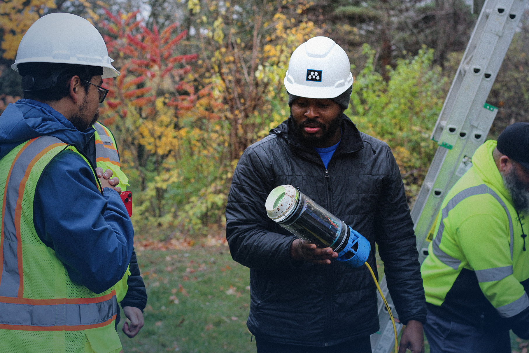 Three outdoor workers wearing safety helmets and jackets, one holding a cylindrical device with a cable, standing near a ladder in a forested area.