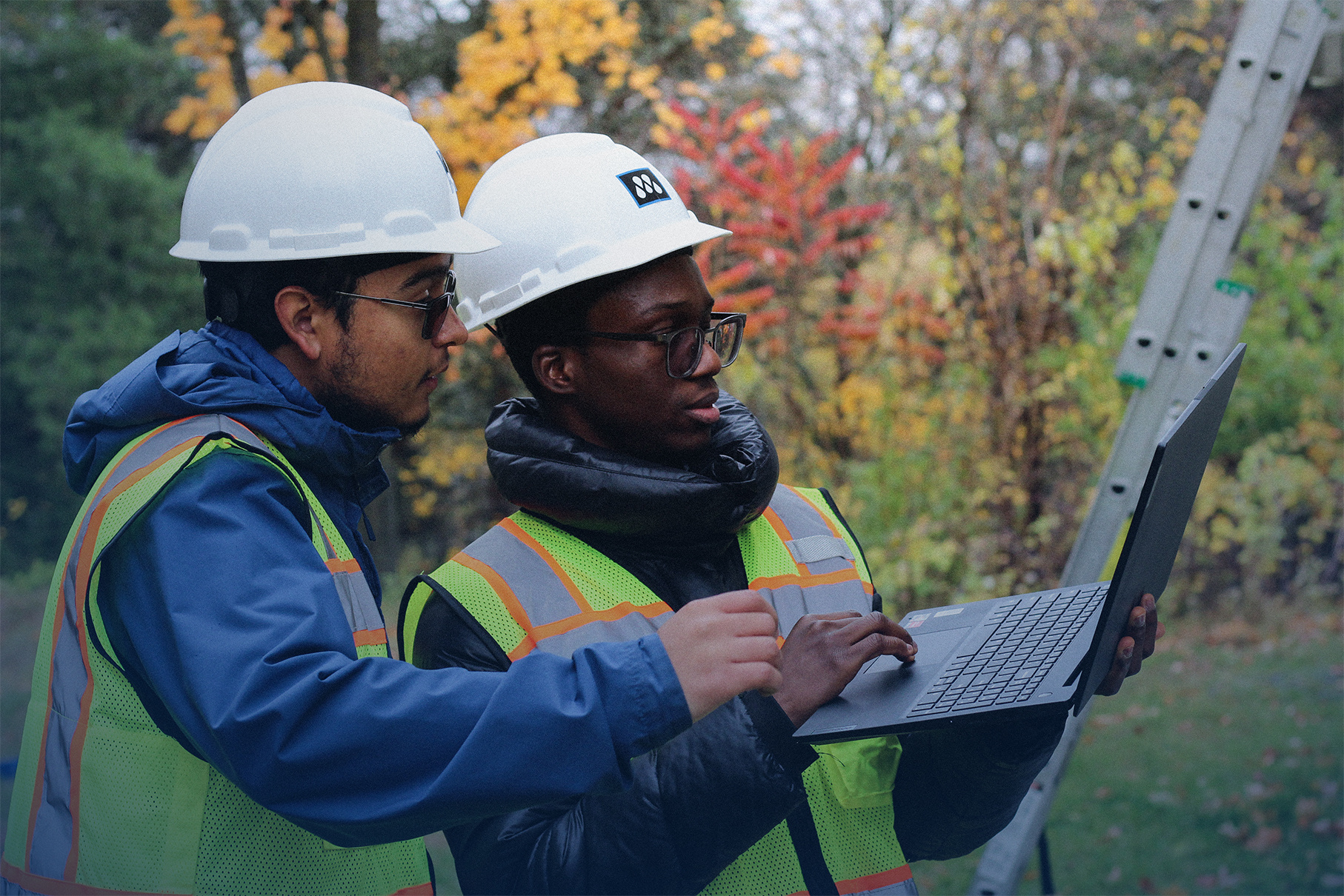 Two construction workers in safety vests and helmets reviewing information on a laptop outdoors.