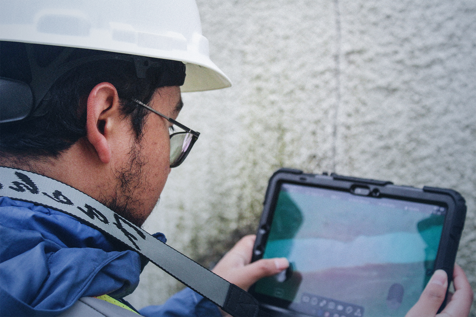 Person wearing a white hard hat and glasses holding a tablet device with a gray strap around their neck.