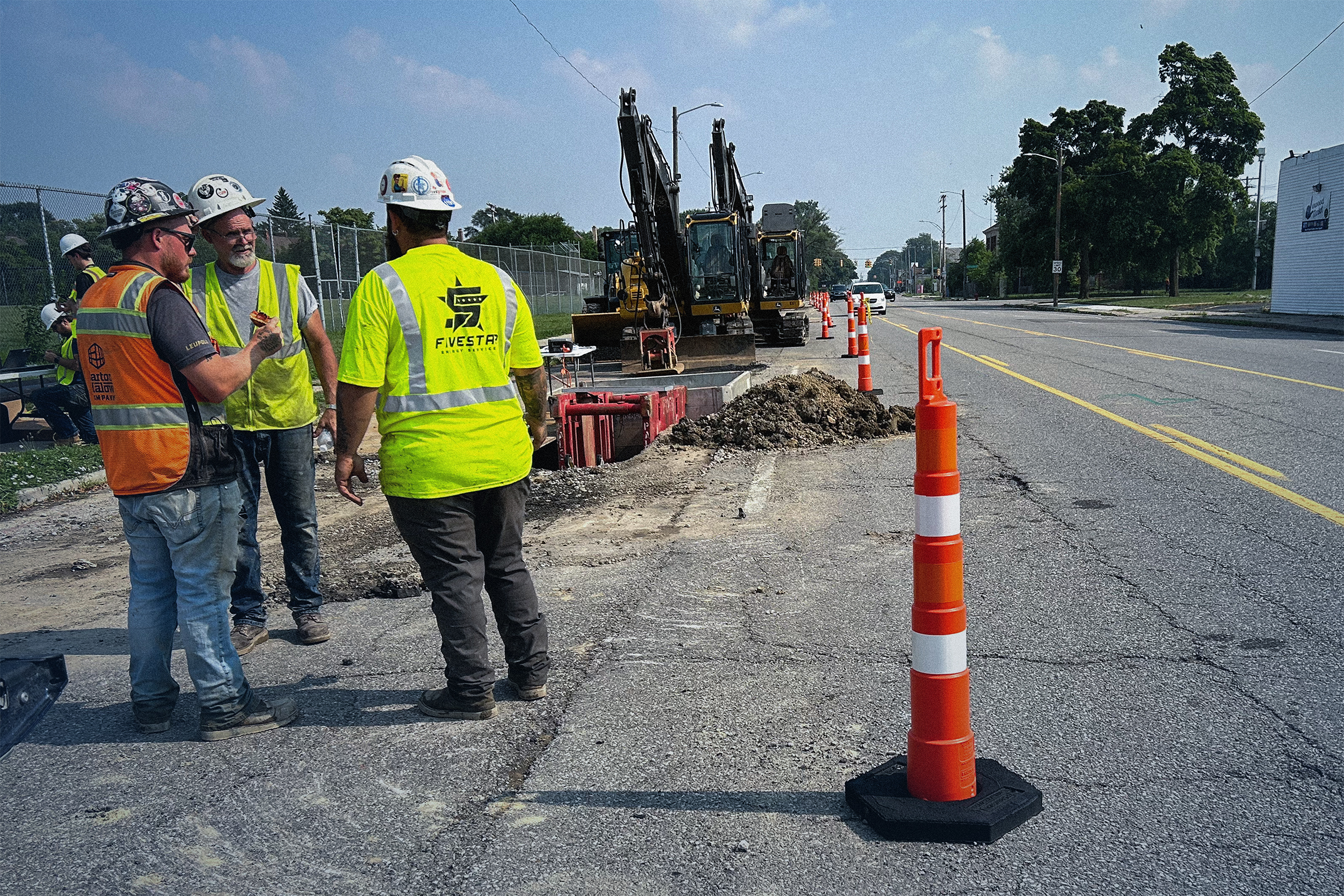 Construction workers in safety vests and helmets gathered near an active roadwork site with excavators and orange traffic cones.