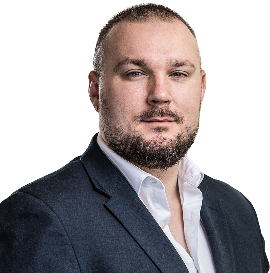 Man with short hair and beard wearing a dark suit jacket and white shirt, looking at the camera.