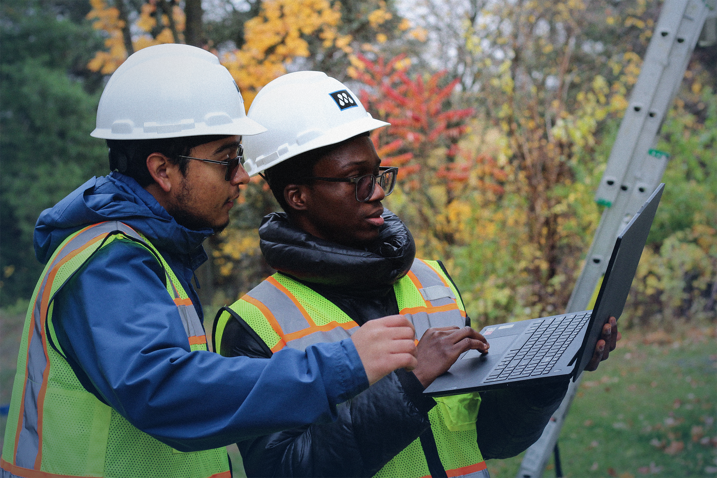 Two construction workers wearing white hard hats and safety vests looking at a laptop outdoors near a ladder.