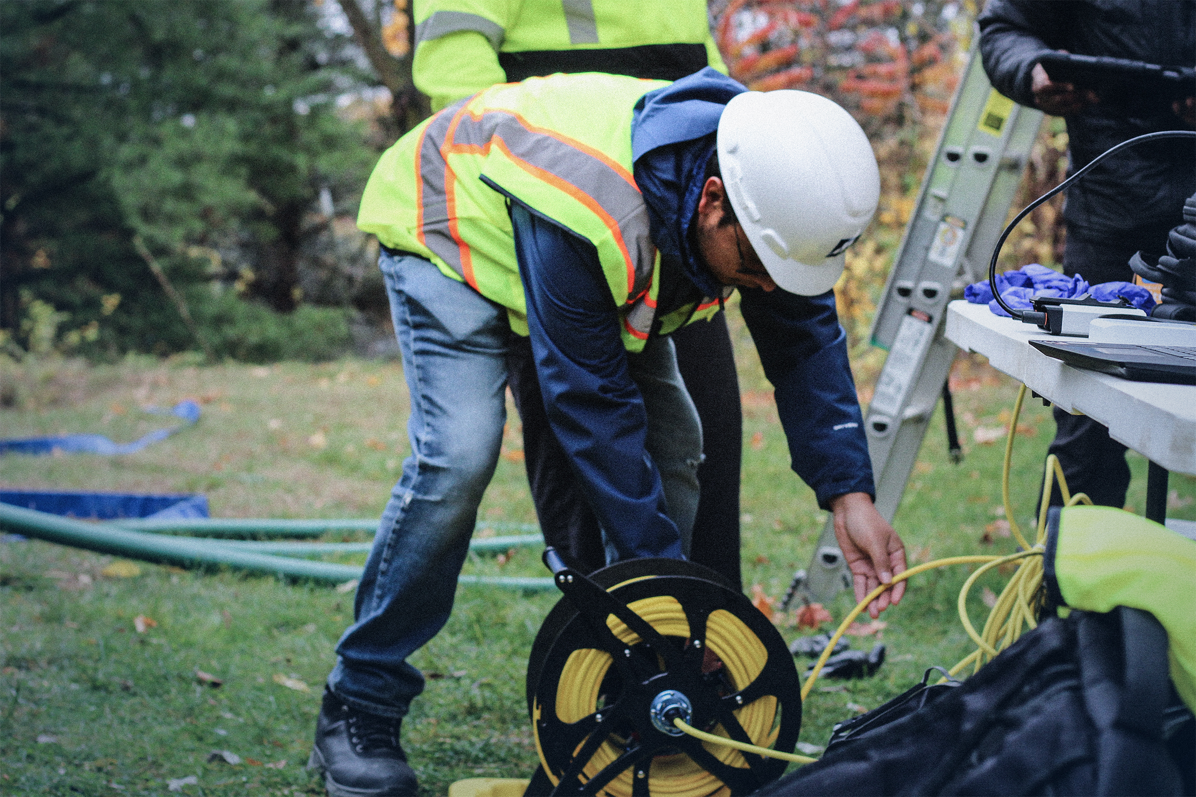 Worker wearing a white hard hat and reflective vest bending down to handle a yellow cable reel outdoors near a ladder and a table with equipment.