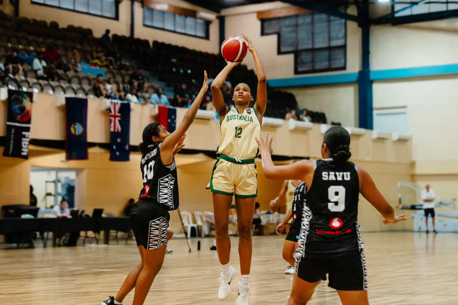Sitaya Fagan in gold rises for a jump shot between two Fiji defenders in black, one contesting with a raised hand, national flags hanging from the balcony behind.
