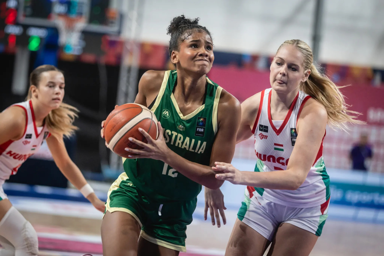 Sitaya Fagan in green drives to the basket with the ball in both hands, braced against a Hungary defender in white who has an arm across her, a second Hungary player trailing behind.