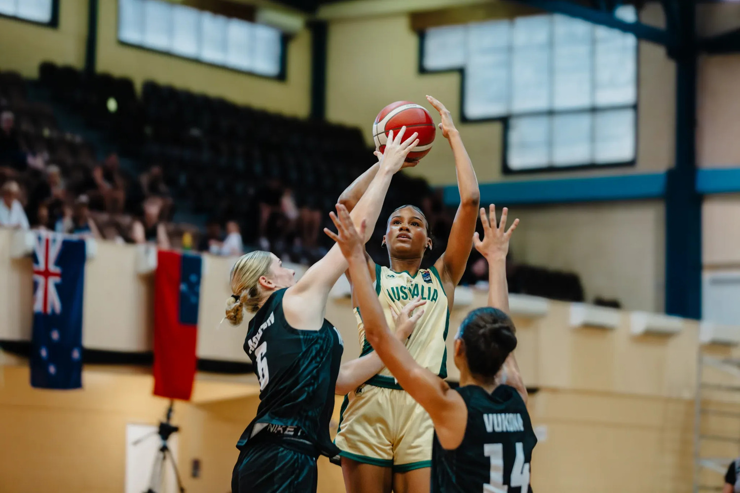 Sitaya Fagan in a pale Australia jersey shoots over two New Zealand defenders in black, both with hands raised, an Australian flag draped on the balcony behind.